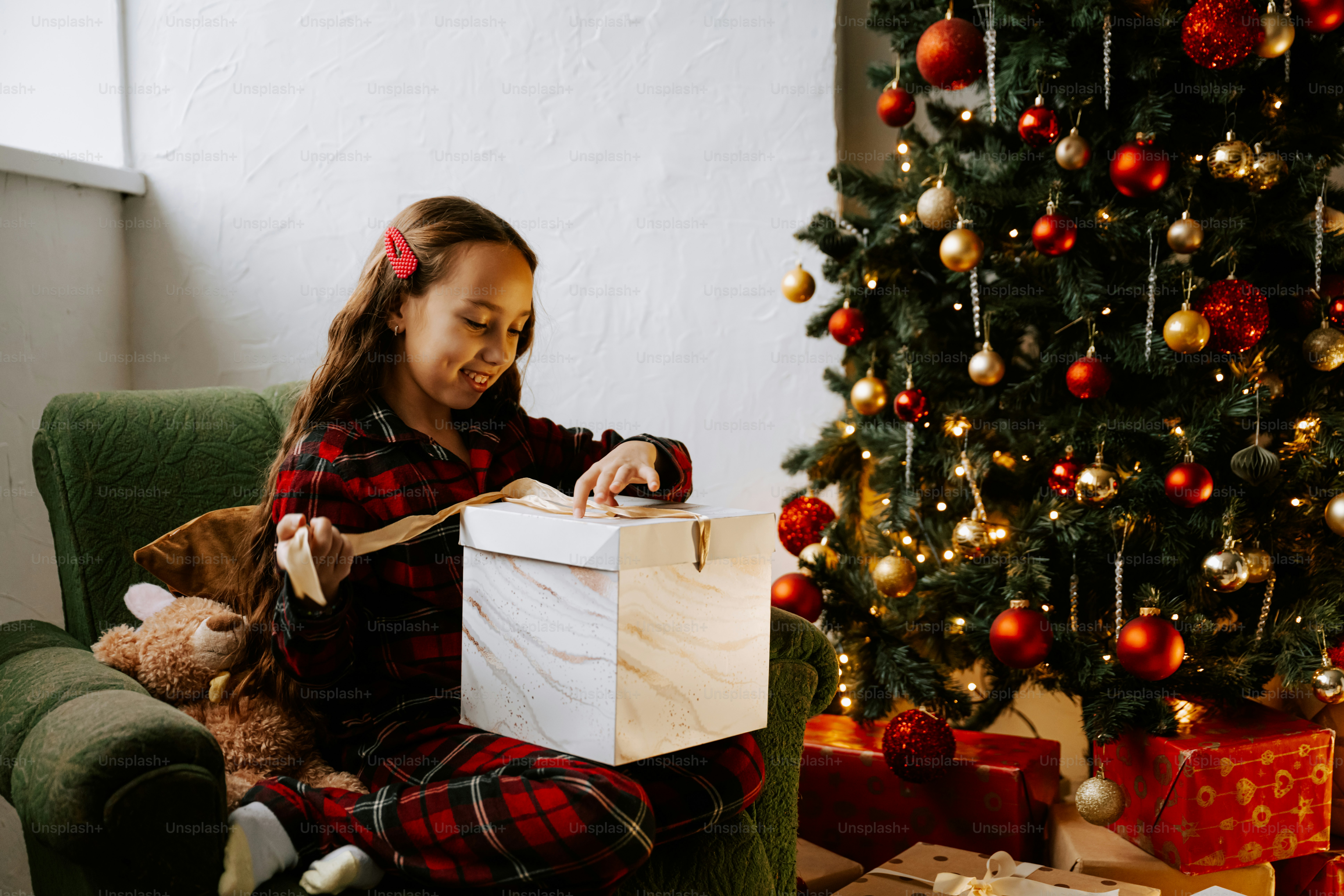 A little girl sitting on a couch with a present in front of a christmas tree