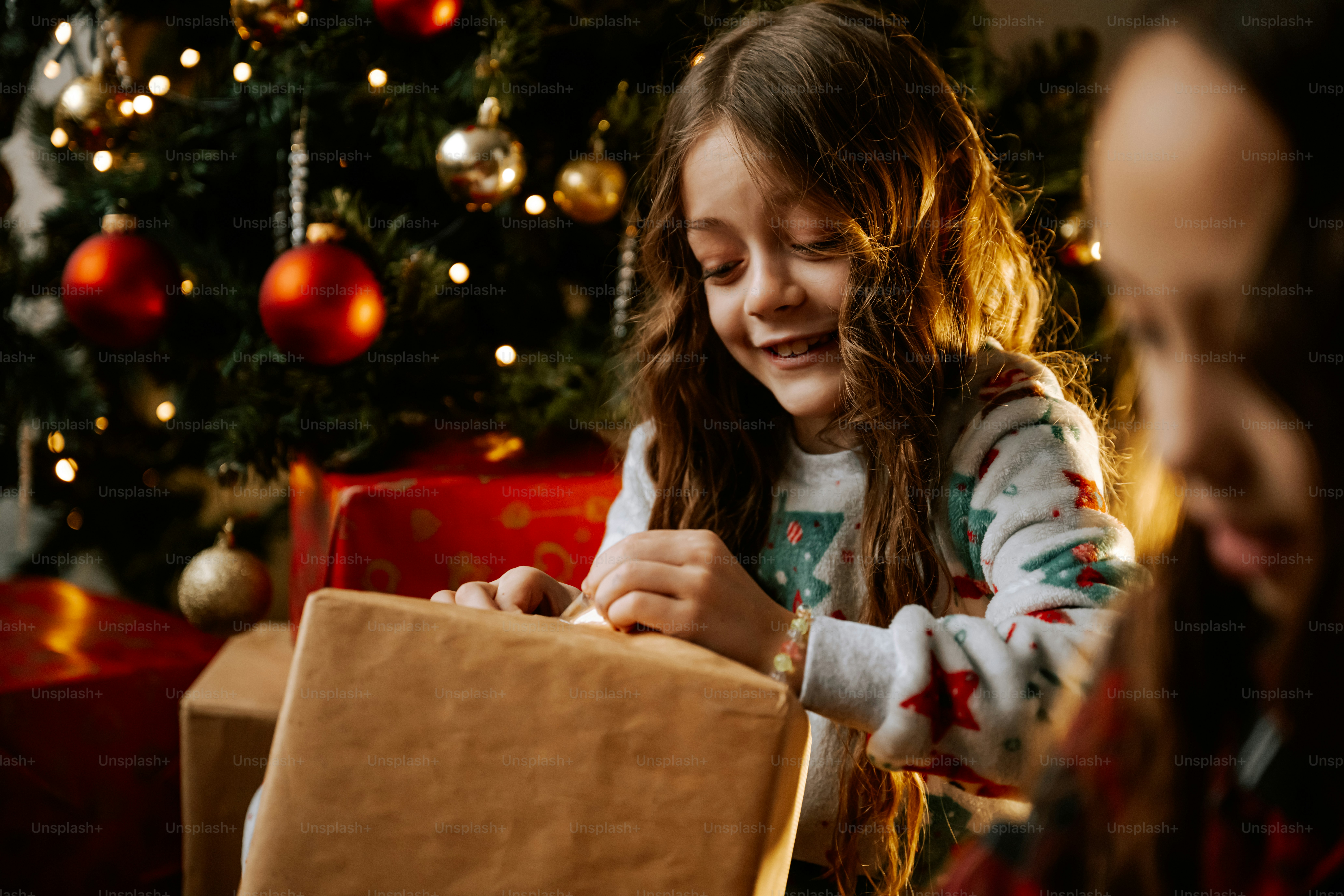 A young girl opening a christmas present in front of a christmas tree