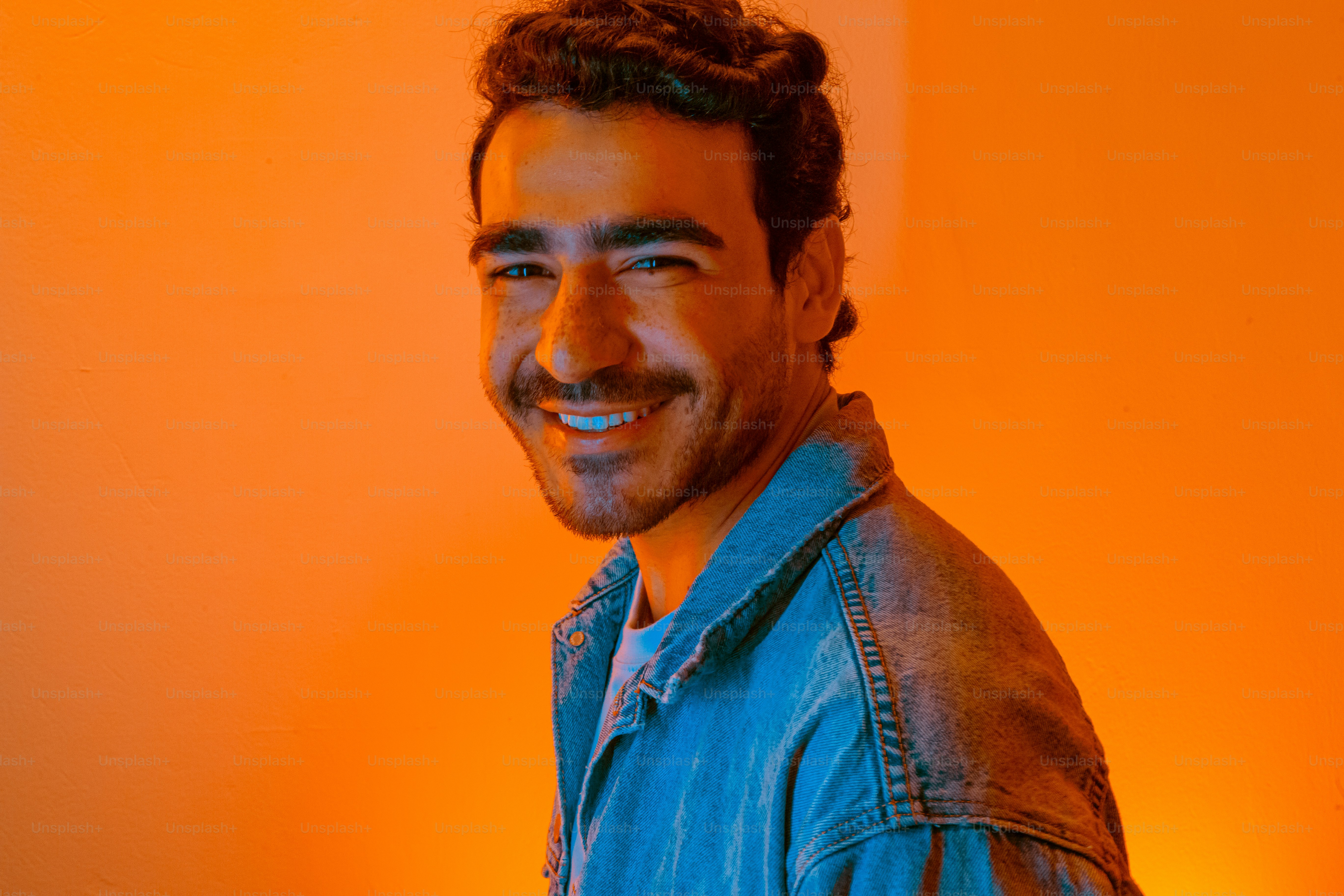 A smiling man in a denim jacket standing in front of an orange wall ...