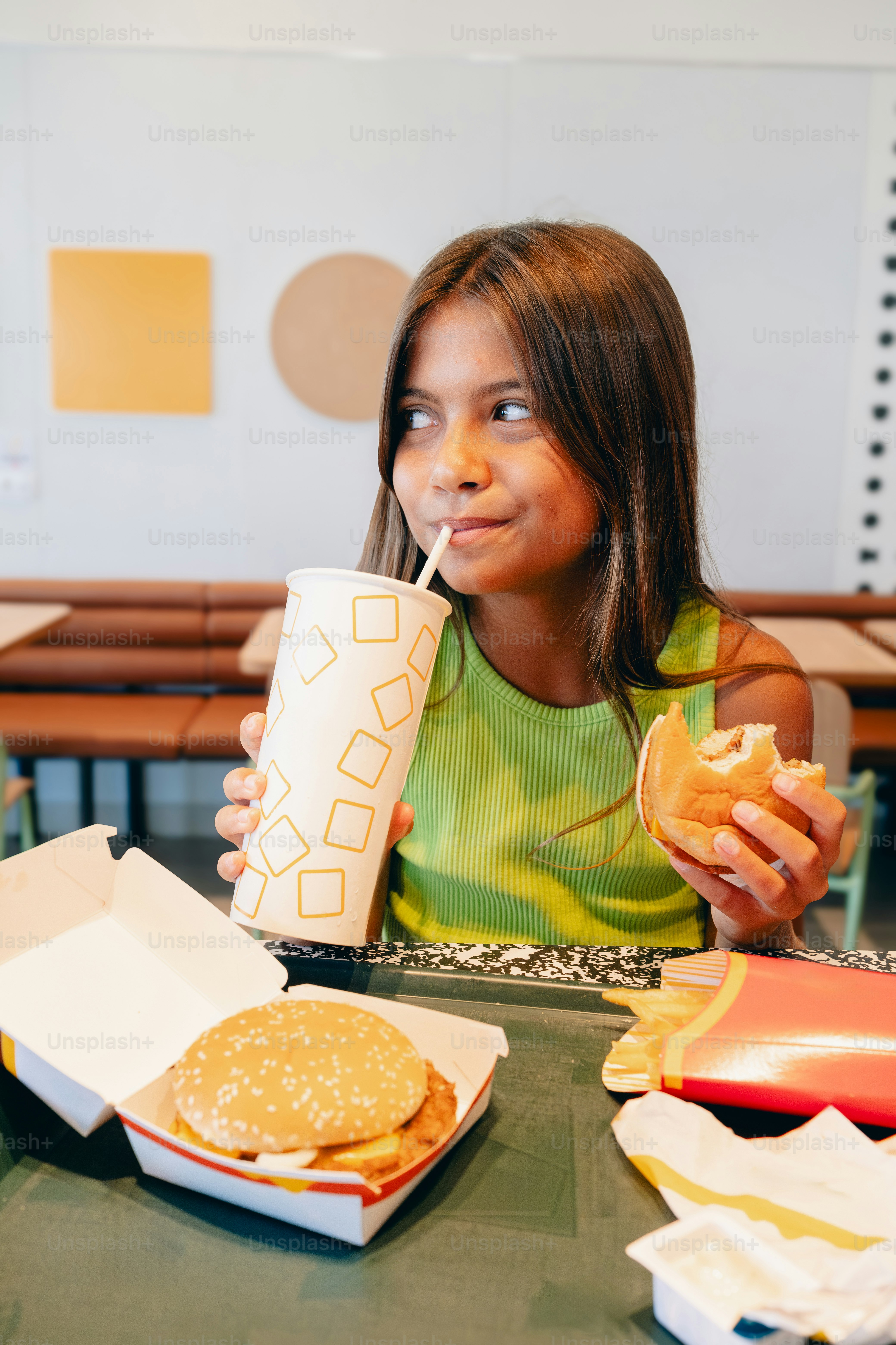 A little girl sitting at a table eating a sandwich and drinking a drink