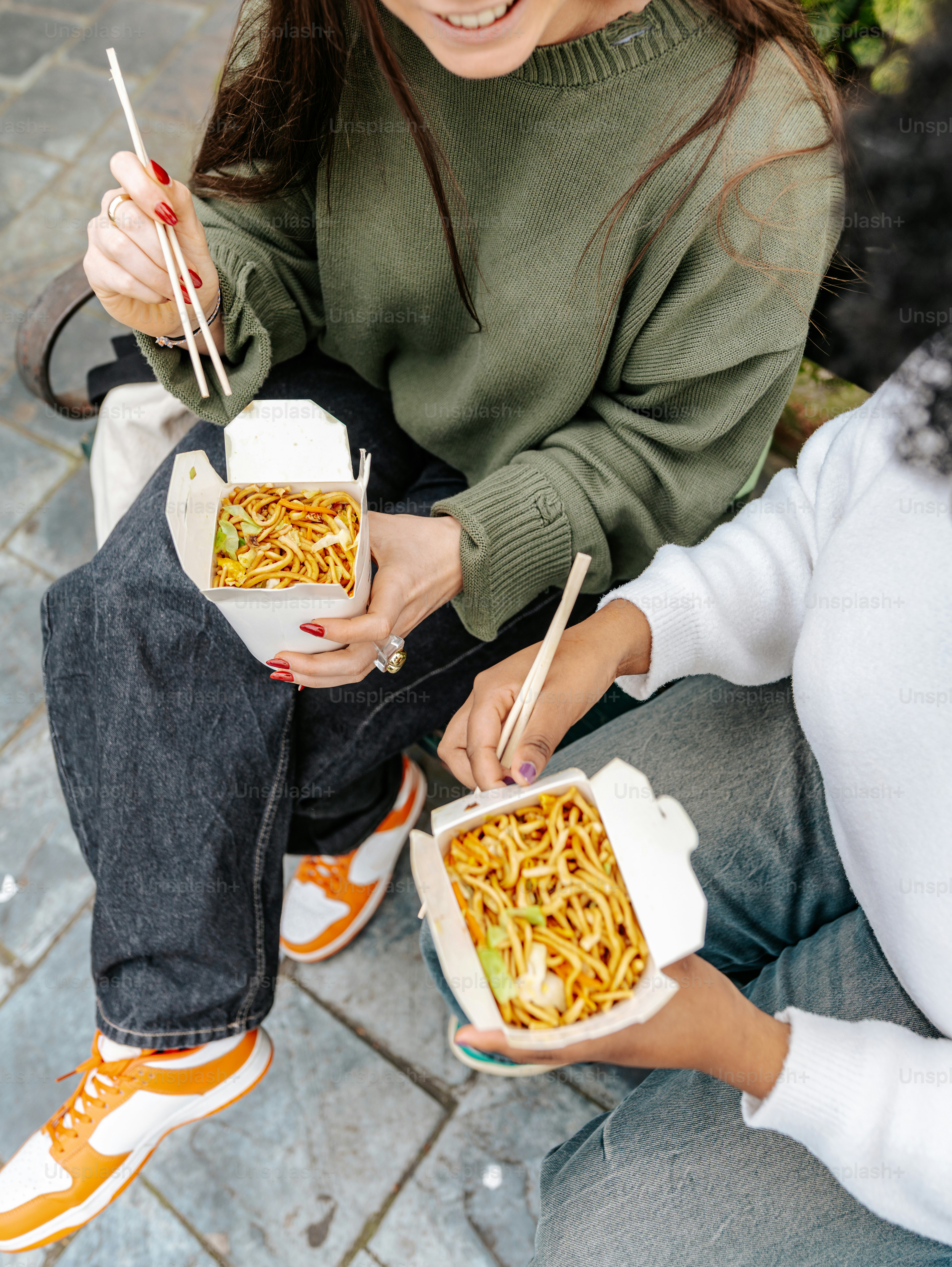 Two women sitting on the ground eating noodles