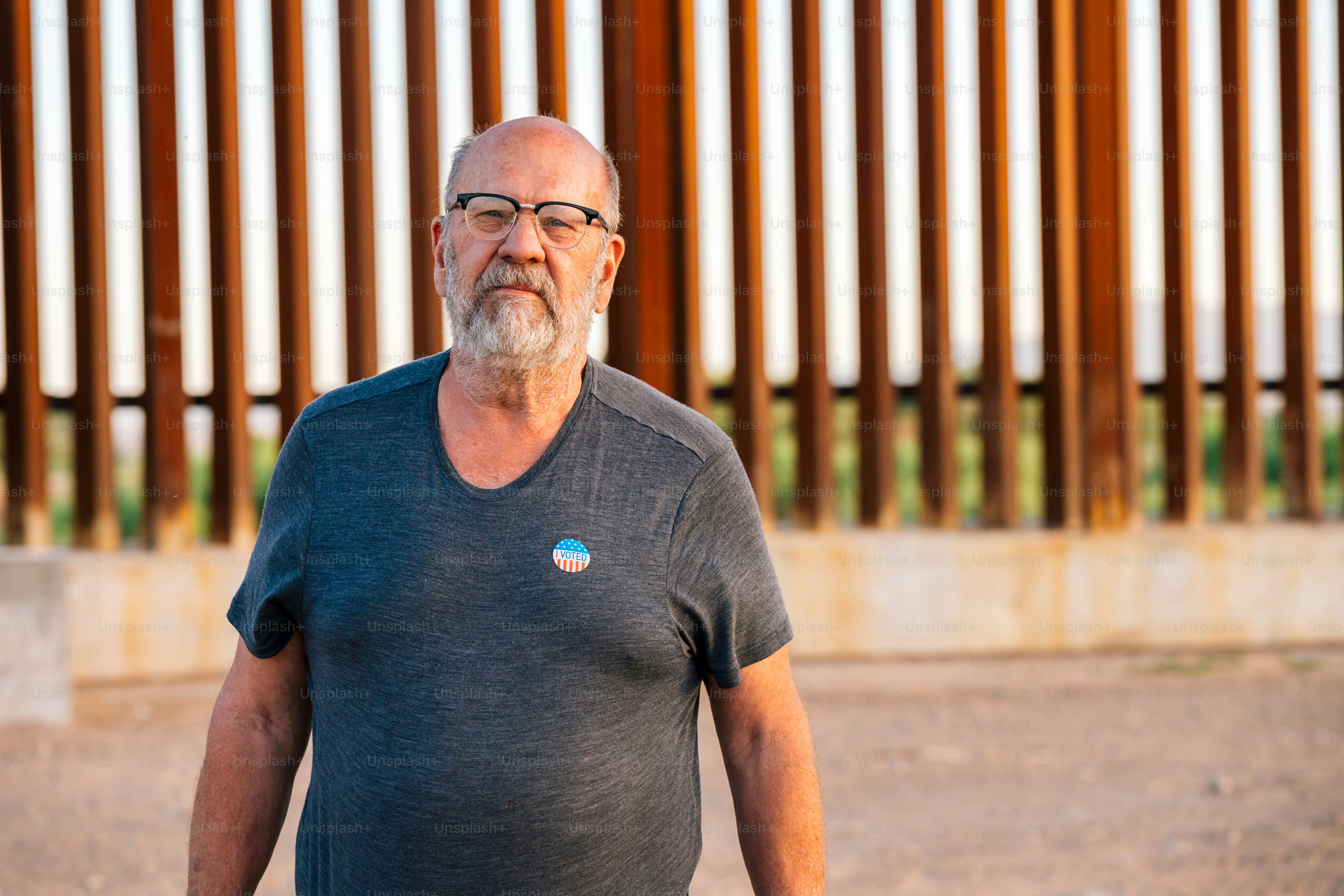 Mature Caucasian Man Wearing an "I Voted" Sticker for a Presidential, Senatorial, Congressional, or local election. He is standing at the U.S. Border Wall Fence separating the United States and Mexico in Springtime. Photographed in Texas, USA.