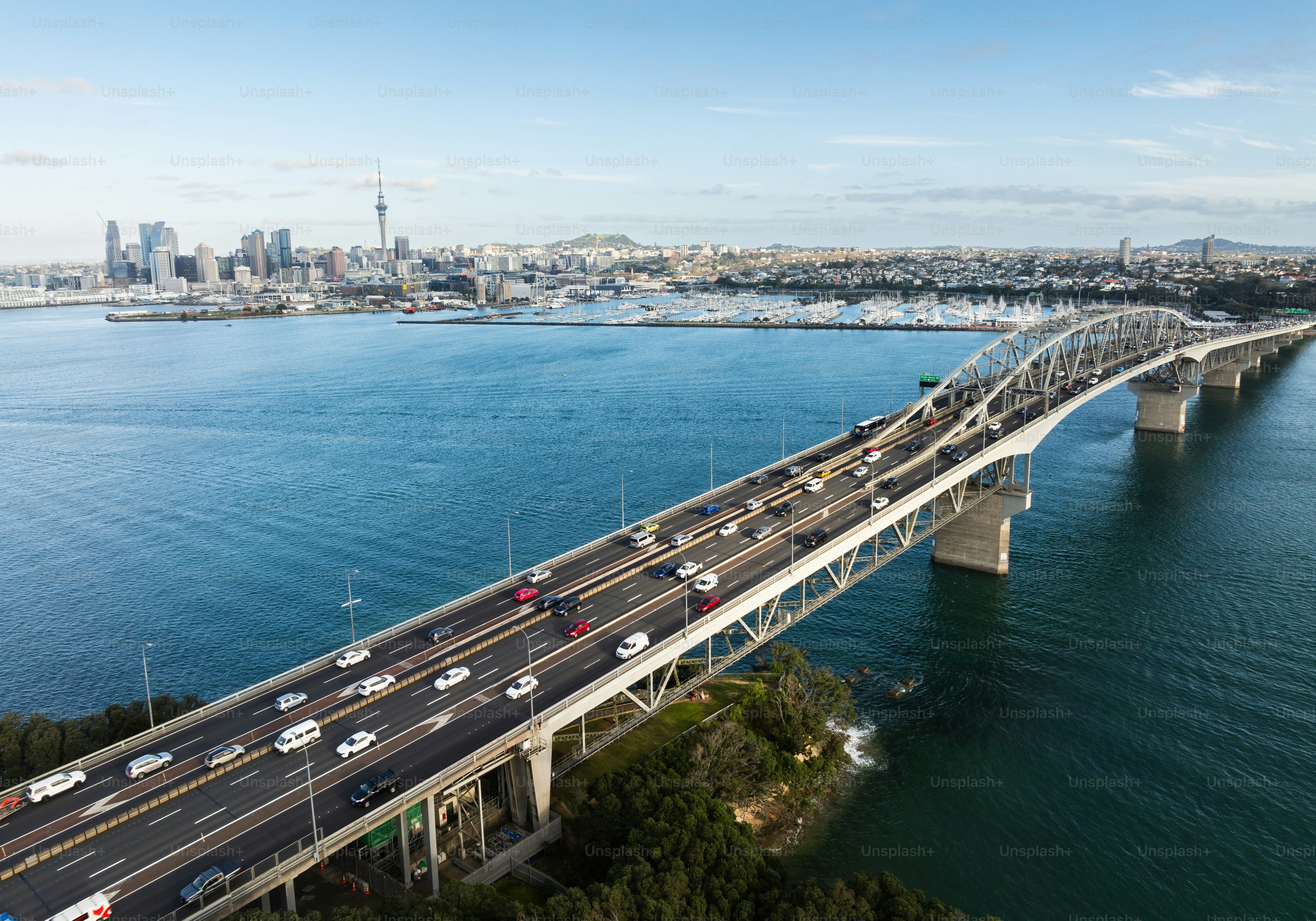 Aerial view of a busy bridge with multiple cars, spanning over a wide blue body of water with Auckland skyline in the background.