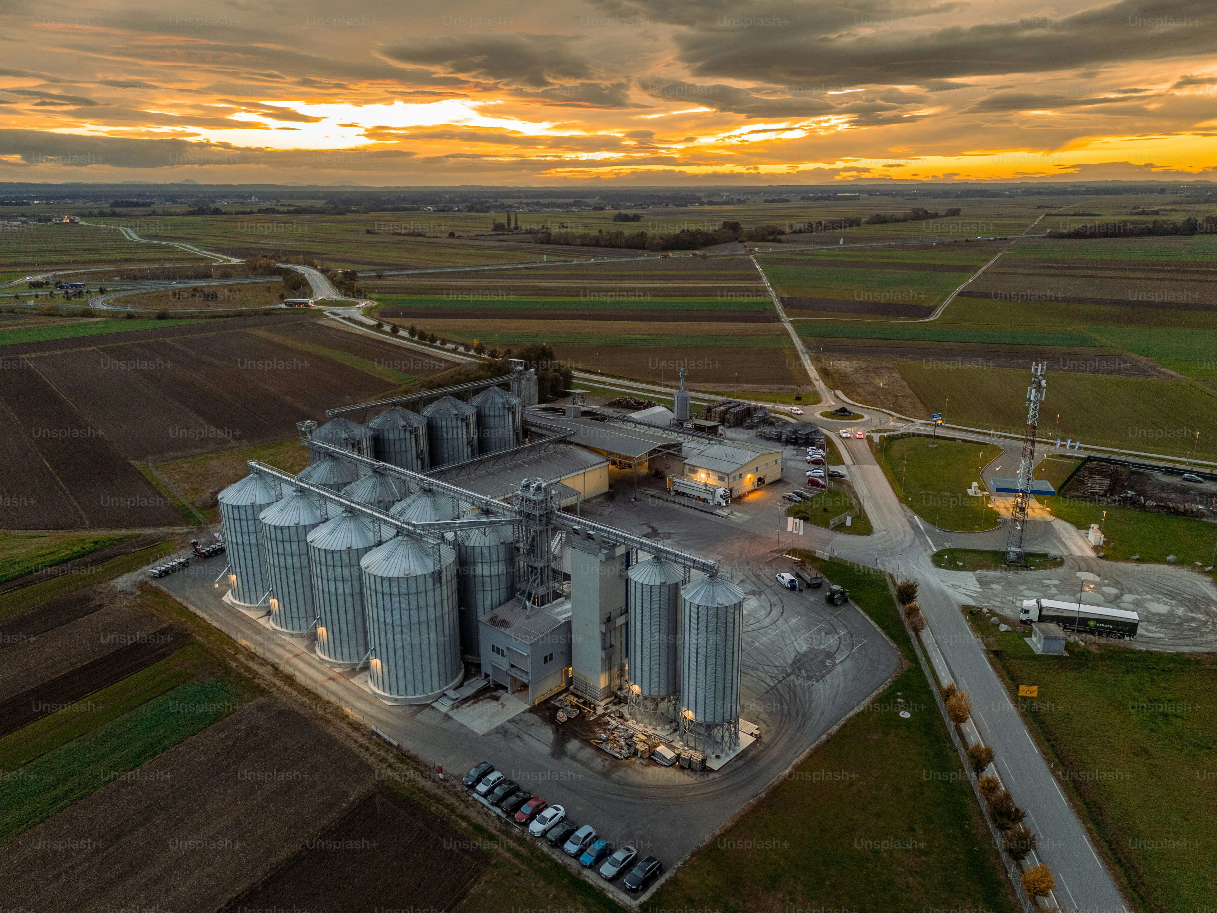 Un vaste champ entoure un grand complexe de silos agricoles sous un ciel de coucher de soleil spectaculaire, soulignant l’importance de la structure pour les opérations agricoles et l’entreposage.