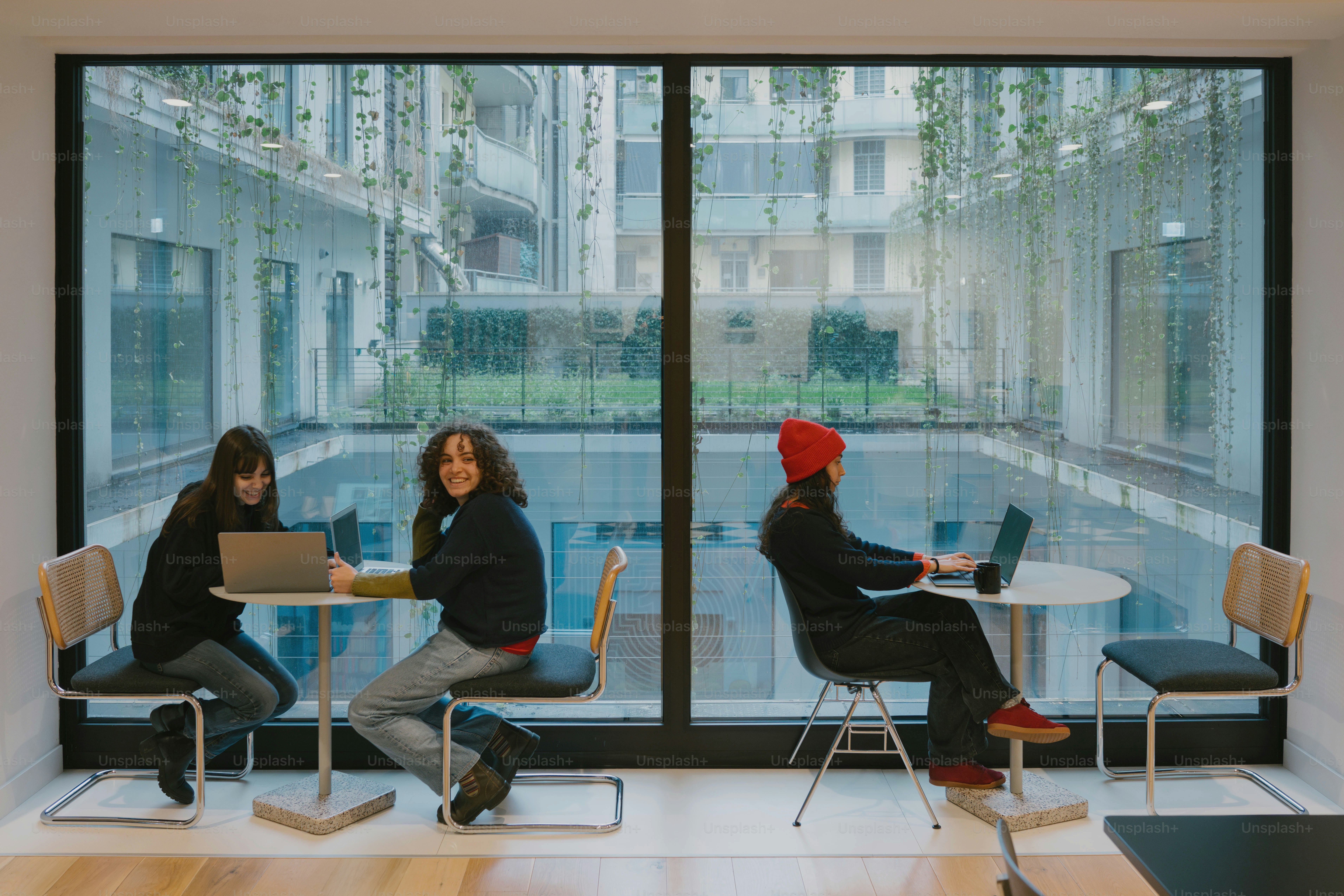 Three people work at tables near a large window.