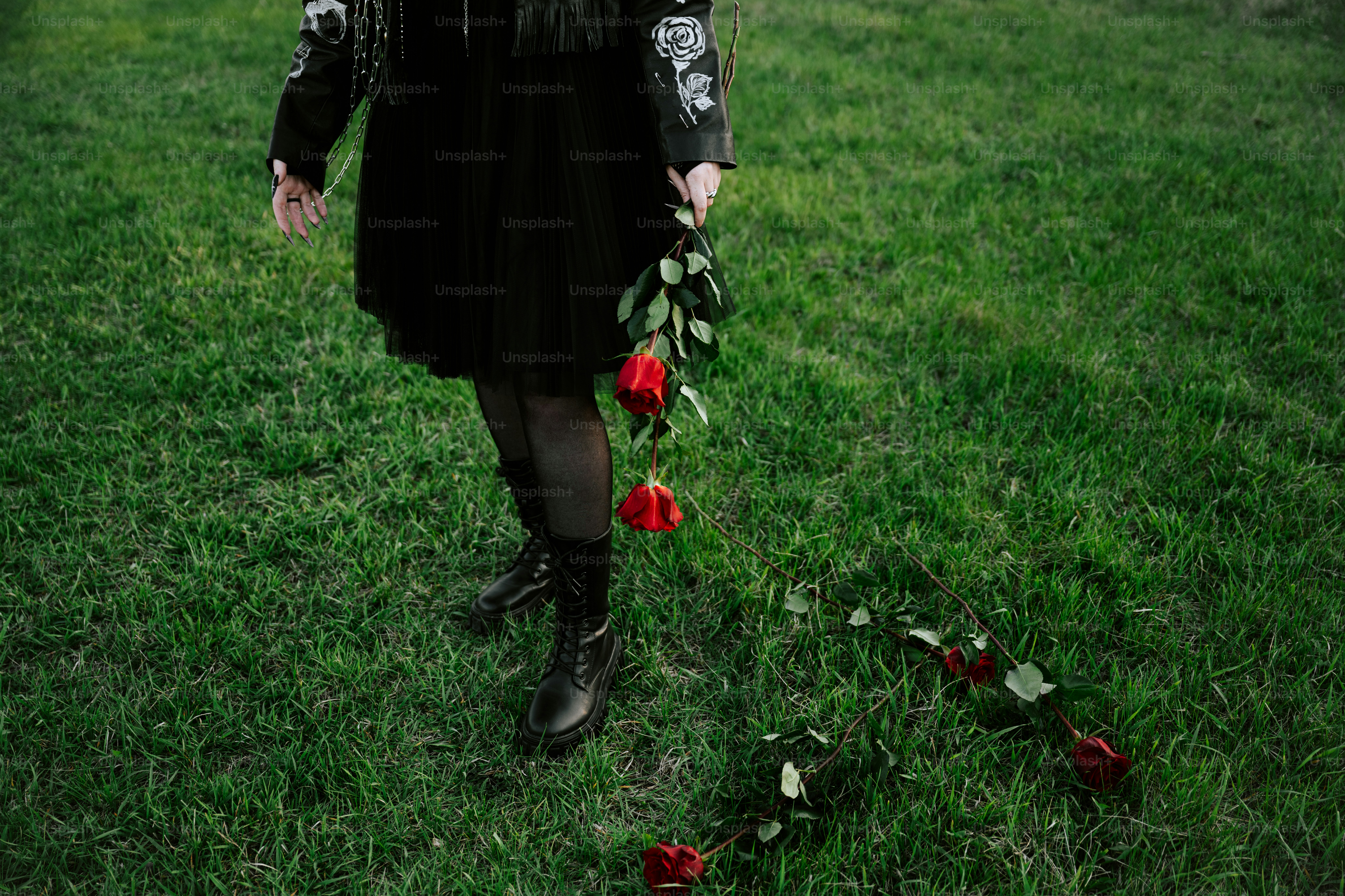 Person in black attire dropping red roses on grass.