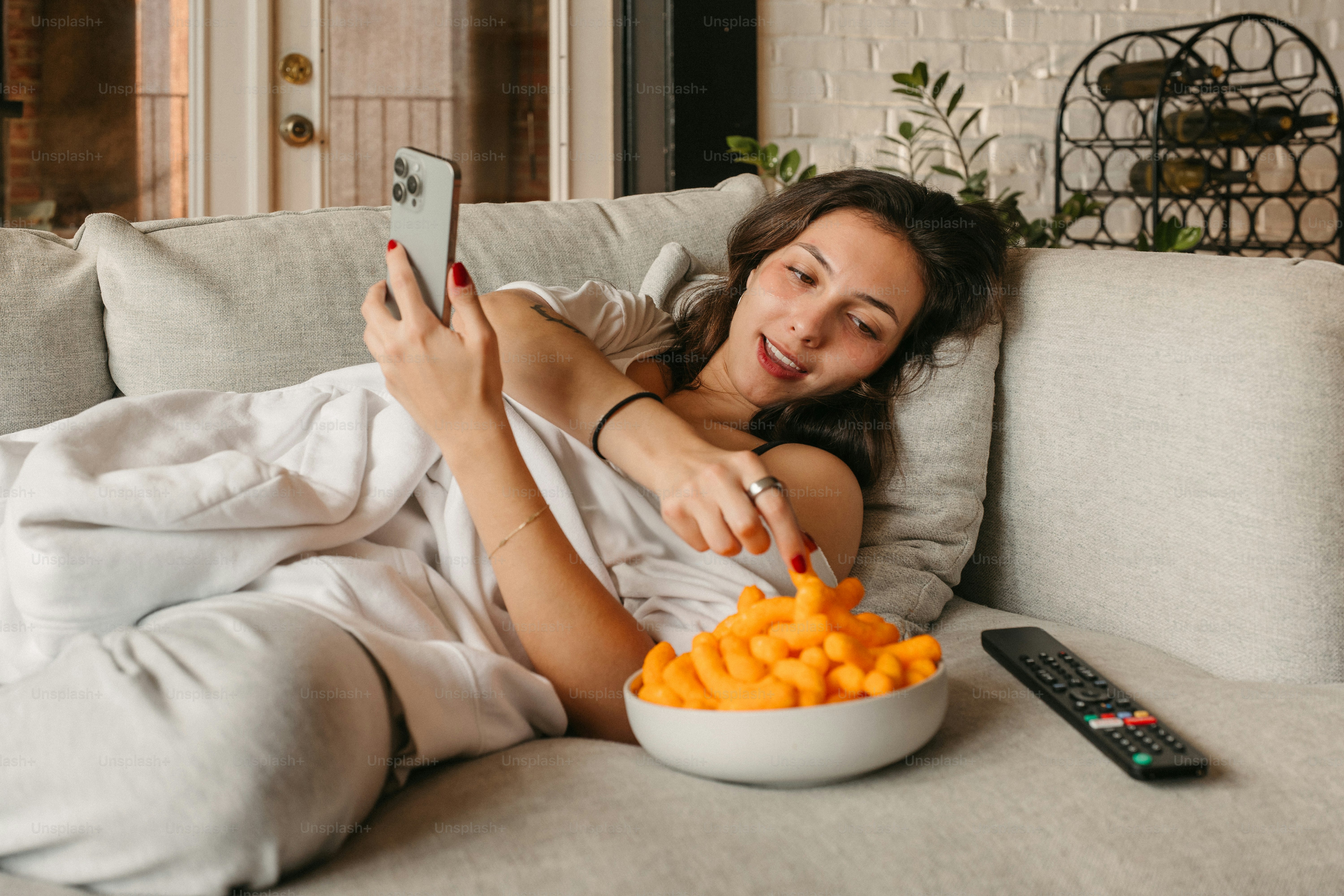 Woman enjoys snacks while taking a selfie on couch.