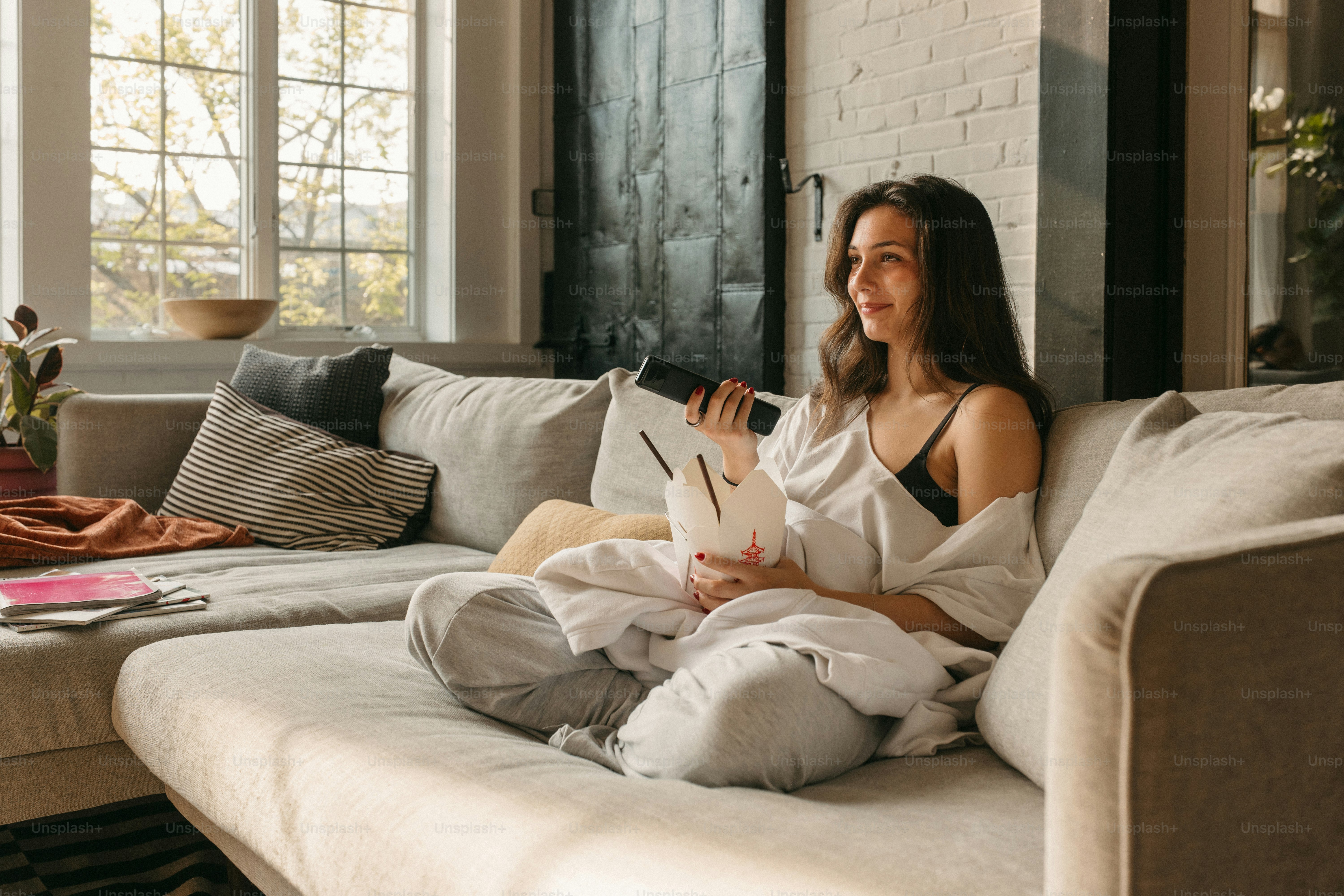 Woman relaxes on couch with takeout and remote.