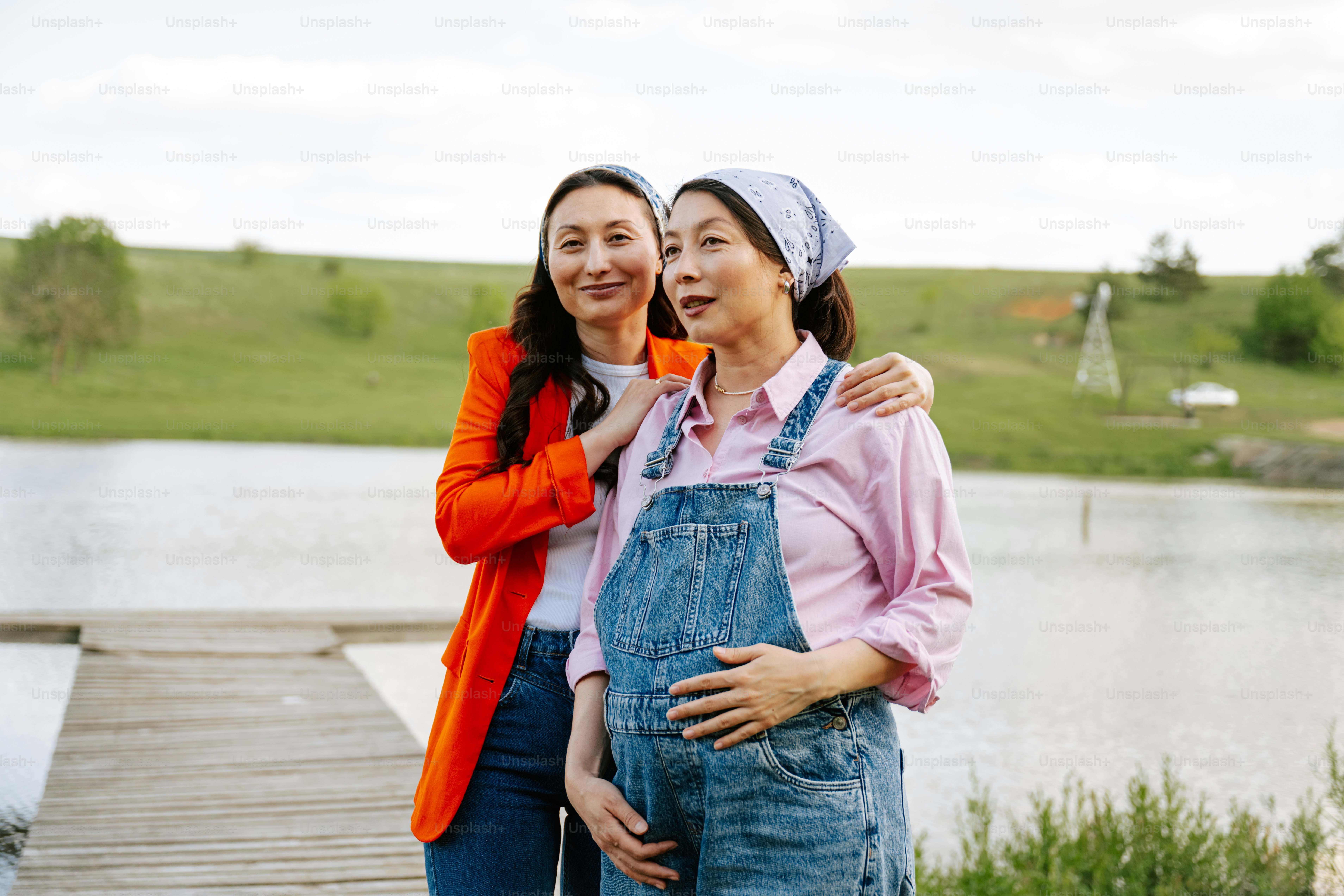 Two women pose by a lake, one is pregnant.