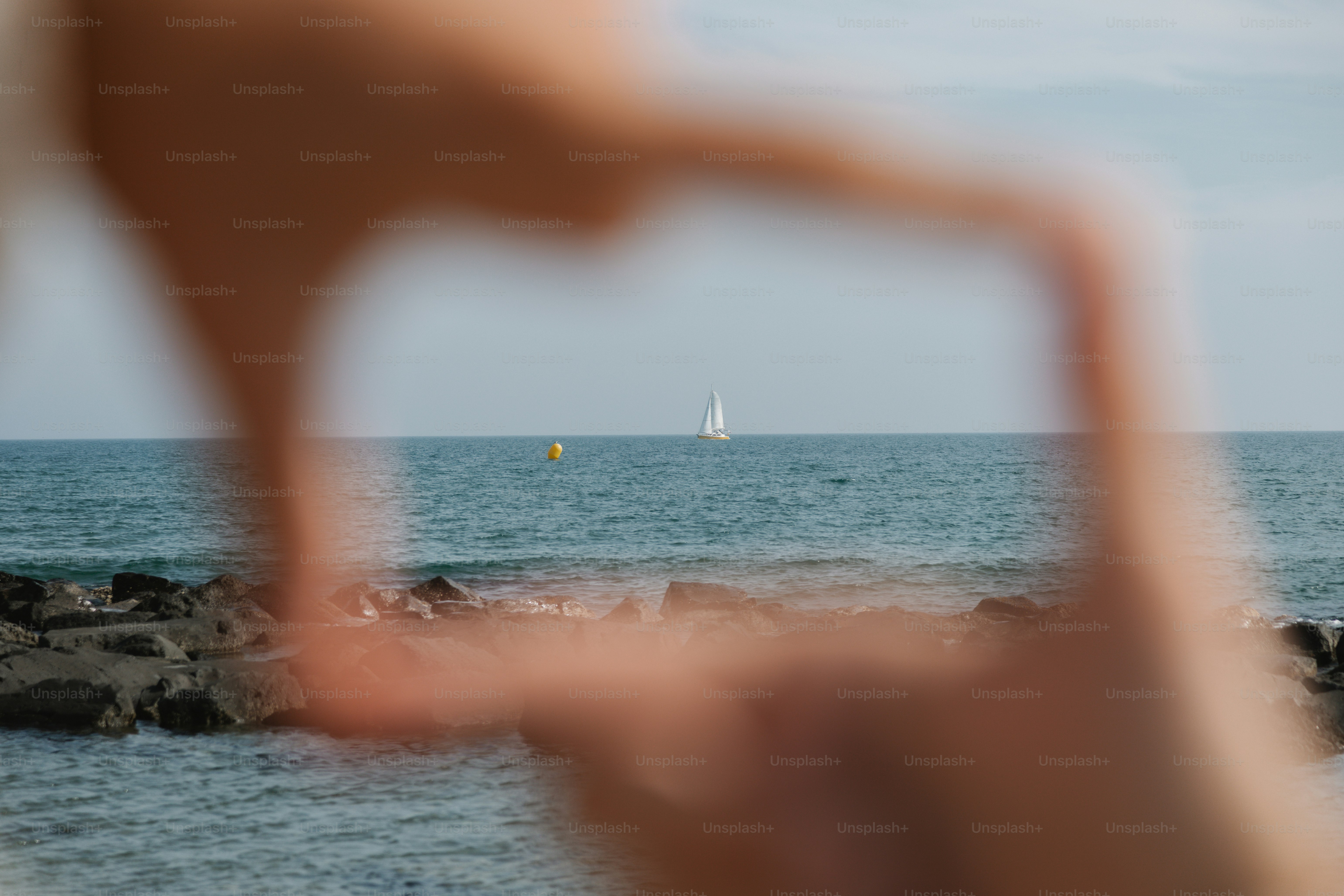Hands frame a sailboat on the ocean.