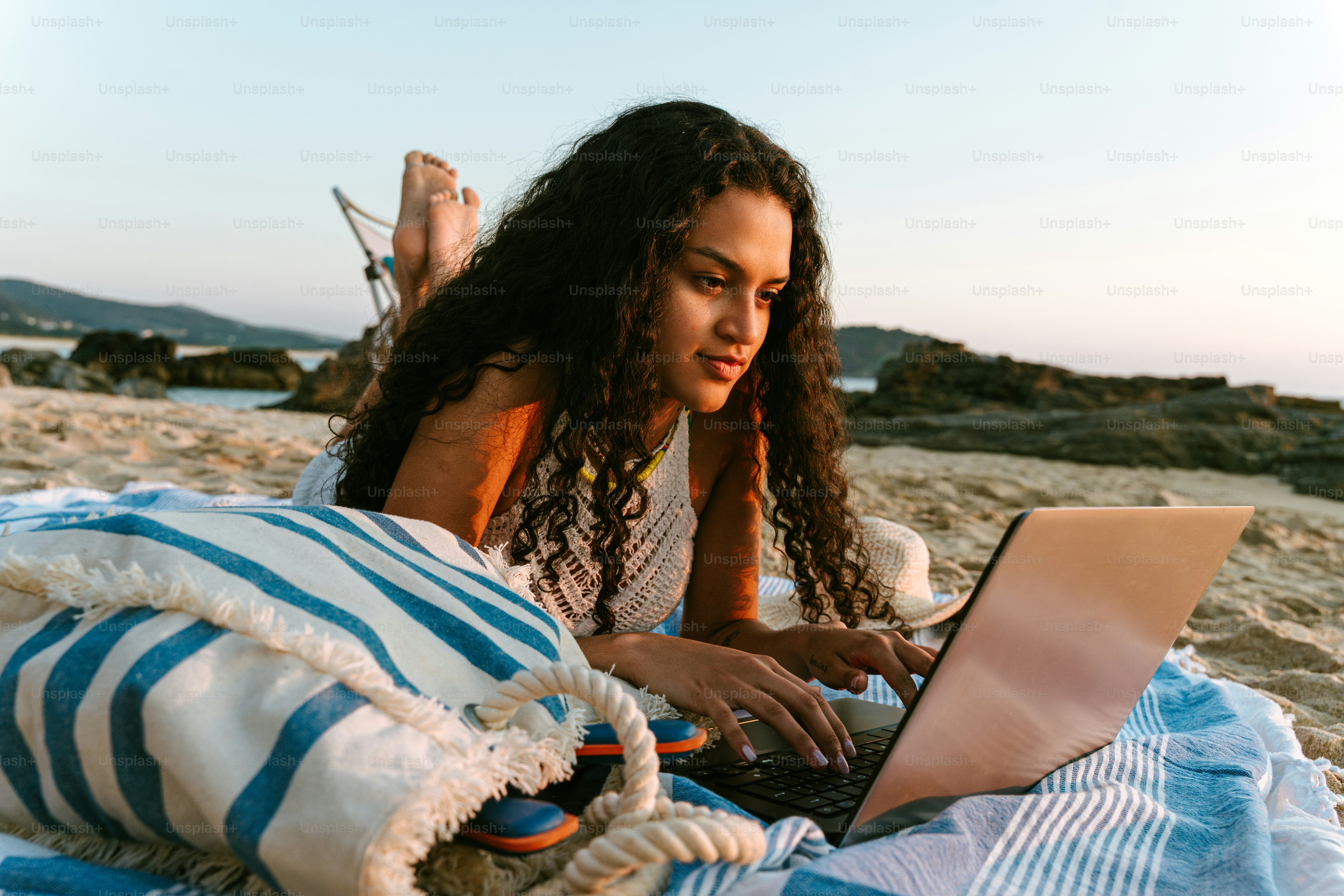 Woman works on laptop while lying on the beach.