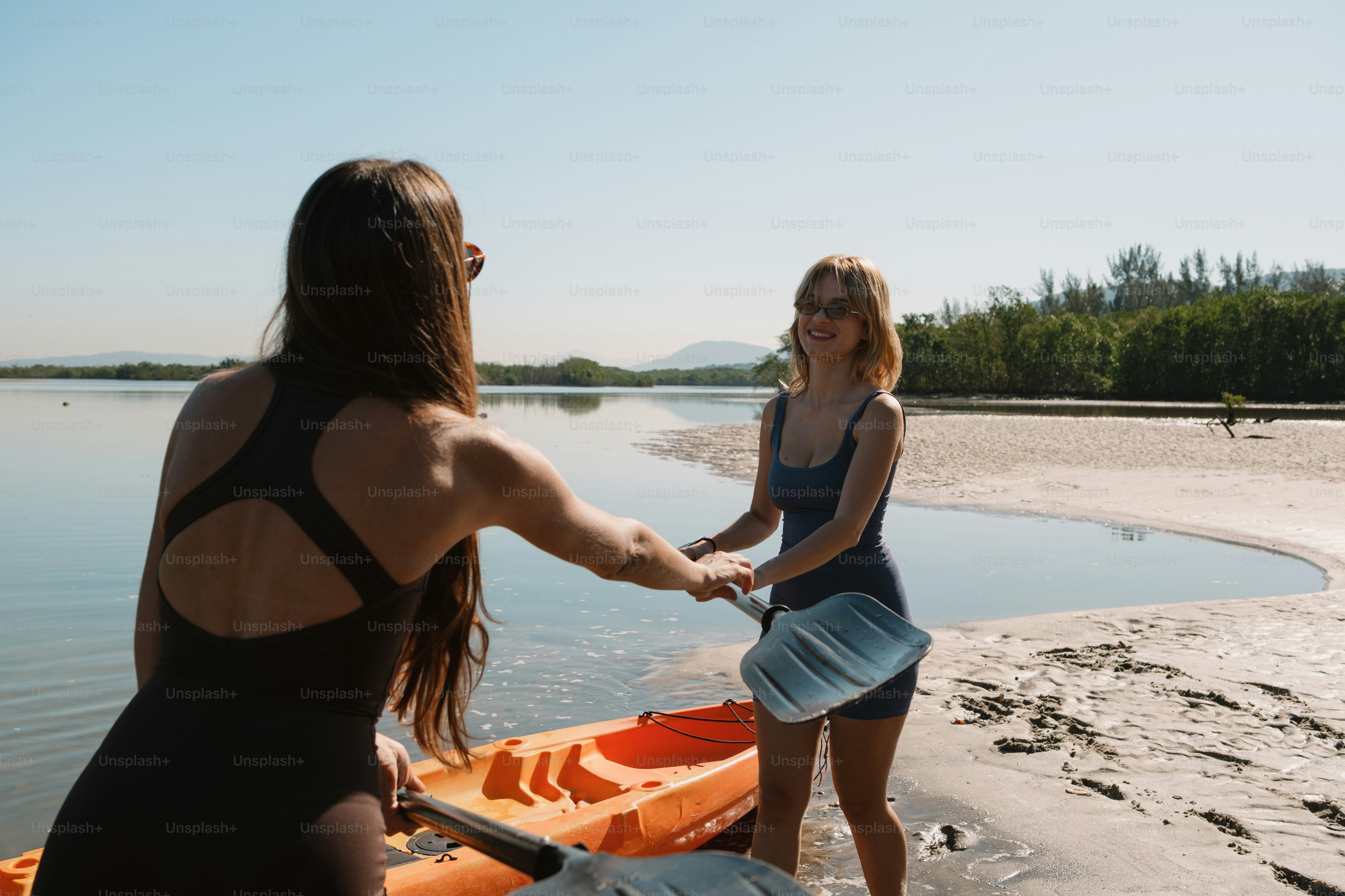 Two women hold hands by the kayaks on the shore.