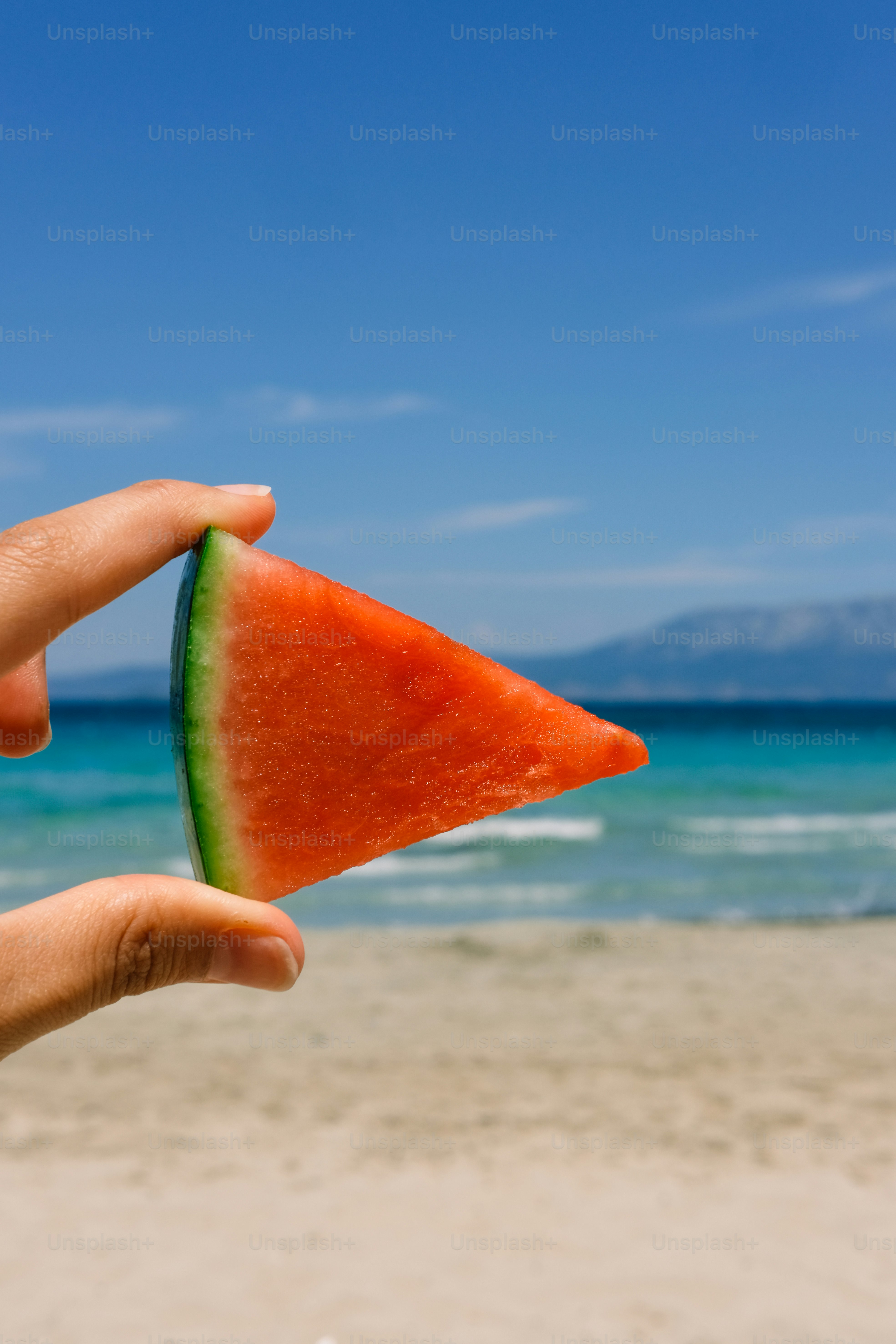 A watermelon slice held up with a beach view.