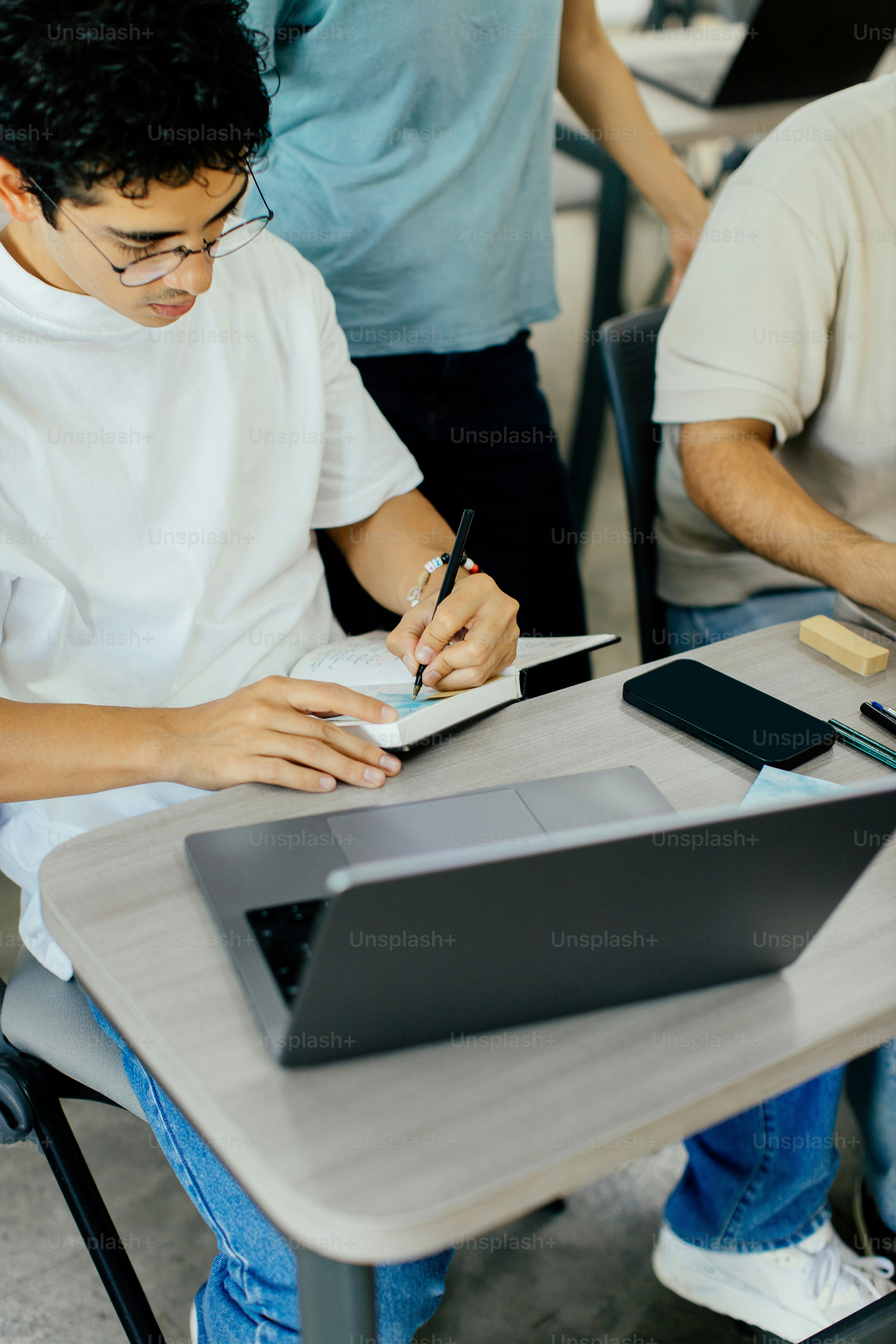 A student taking notes while seated at a desk with a laptop and classroom materials