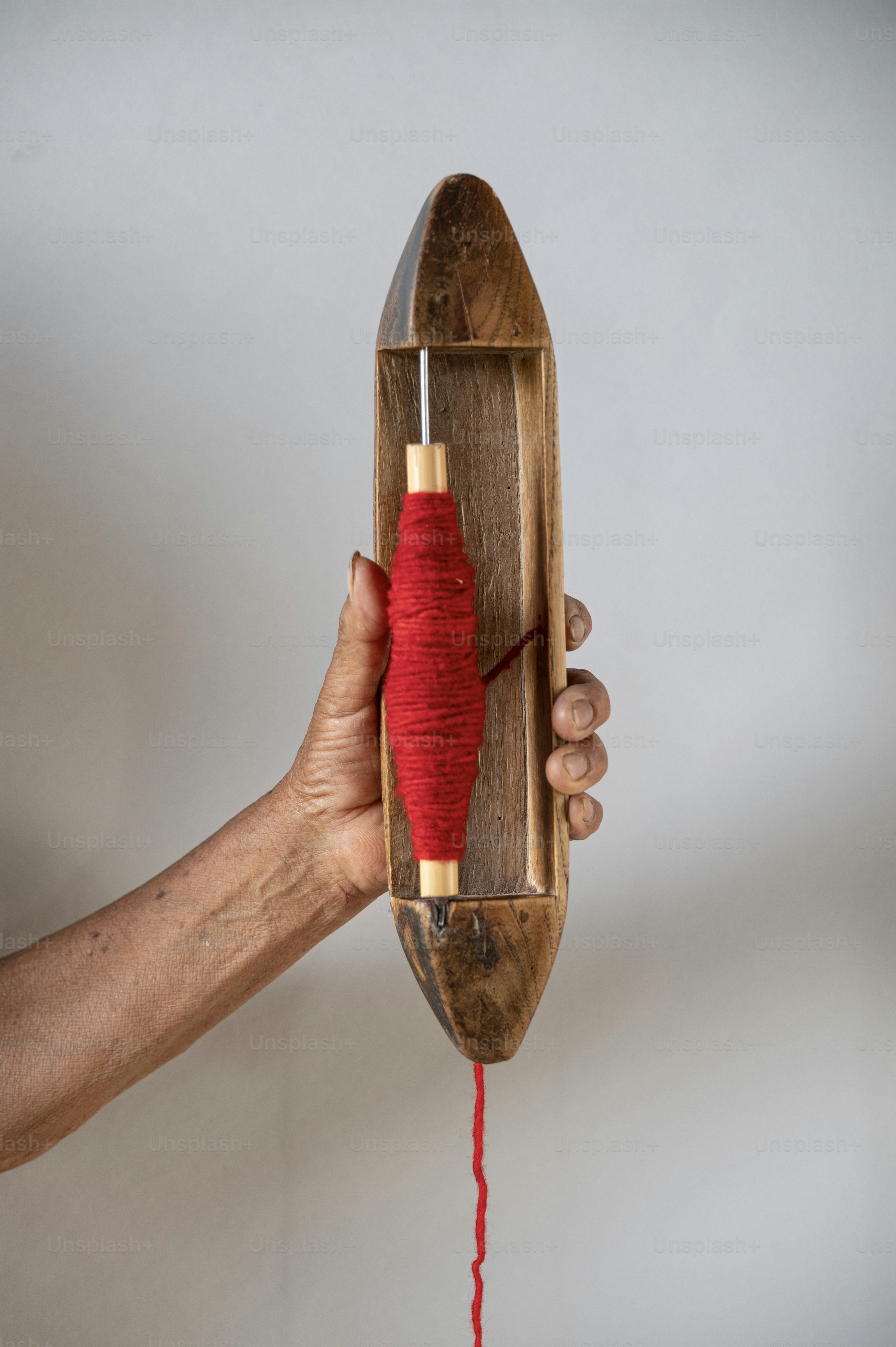 A hand holds a wooden shuttle with red thread.