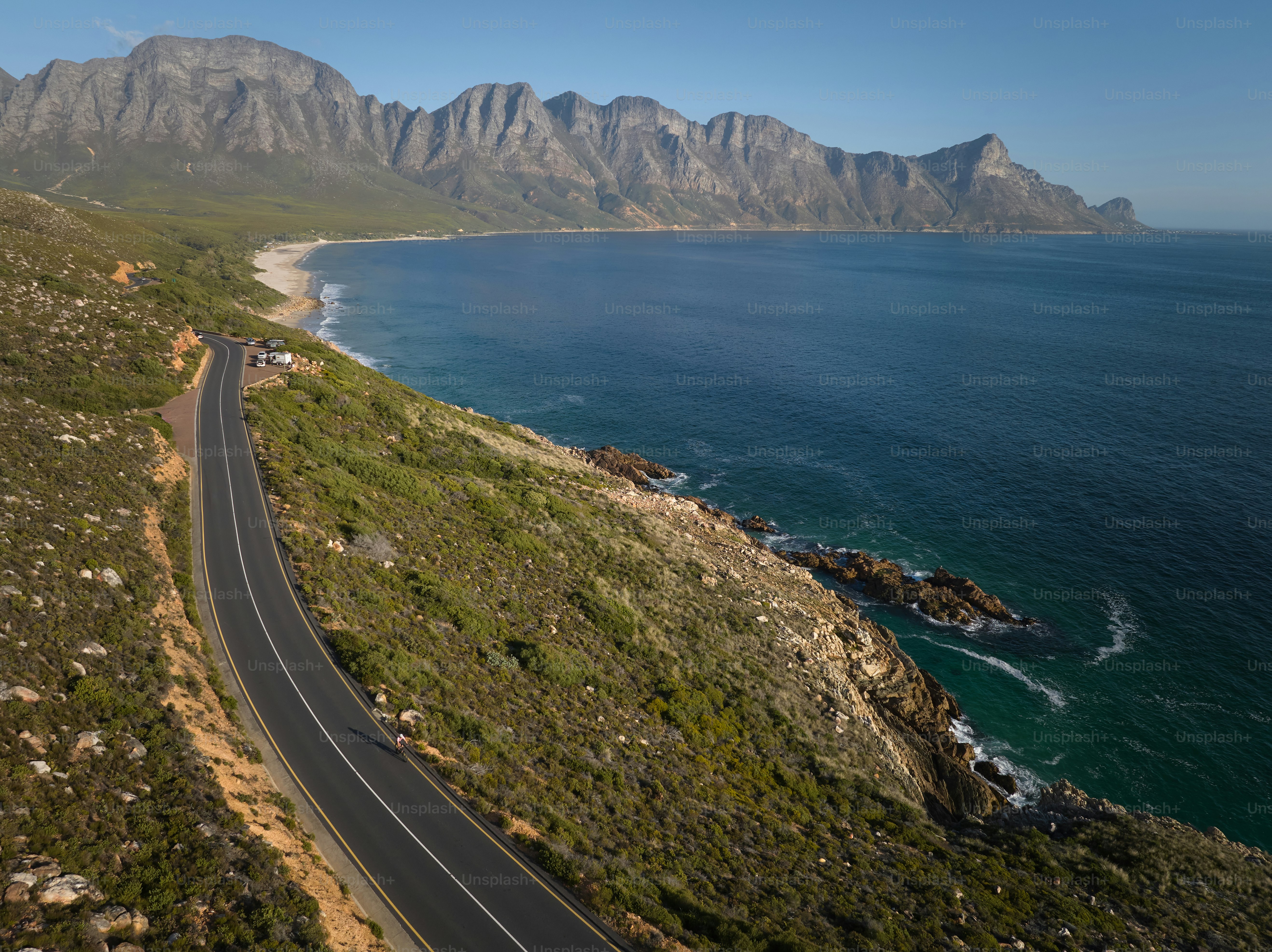 Vue aérienne d’une cycliste chevauchant le long d’une route côtière pittoresque avec l’océan et les montagnes. L’atmosphère tranquille met en valeur la beauté des paysages côtiers et évoque des sentiments de liberté et d’exploration.