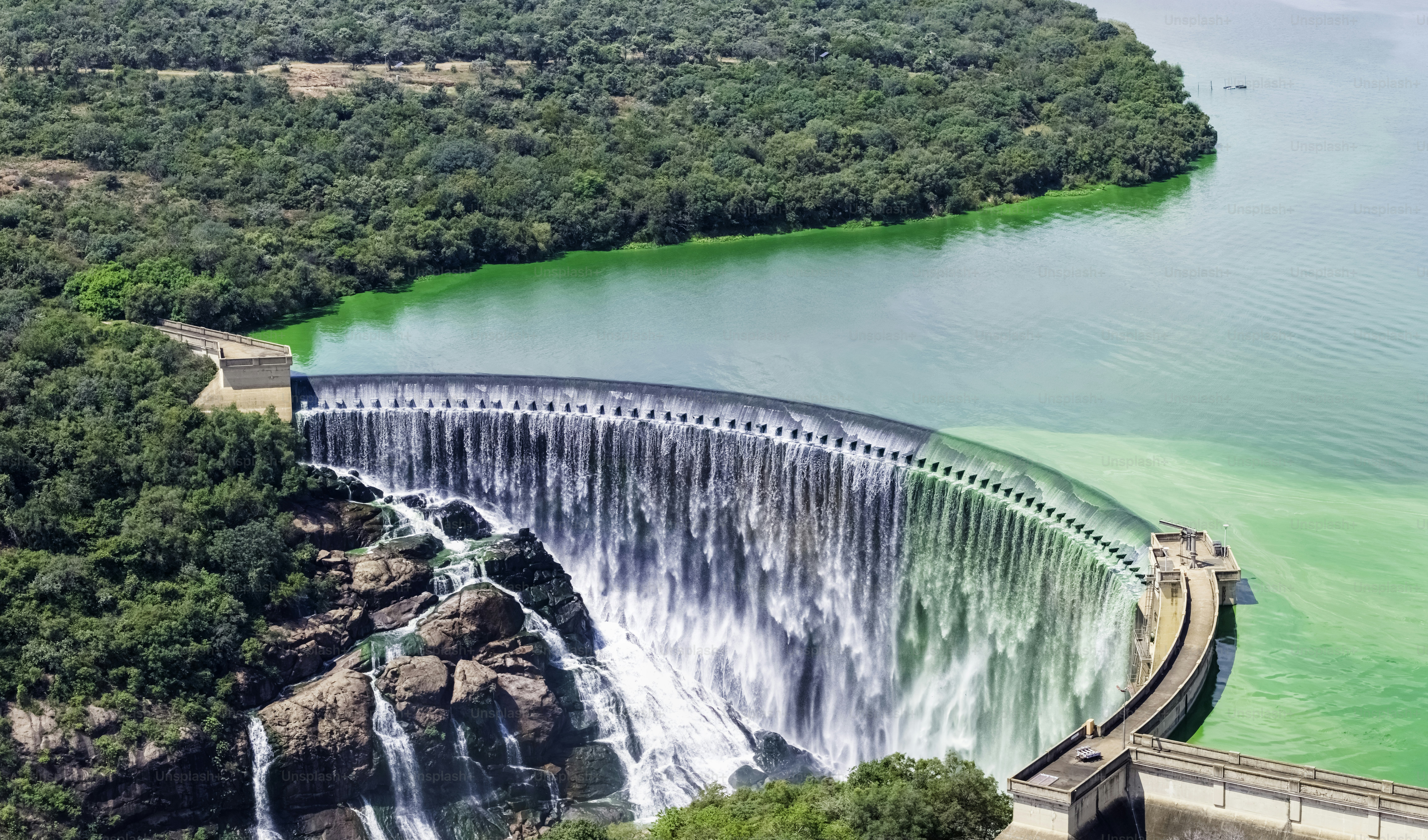 An aerial view of a curved dam overflowing into a rocky river valley