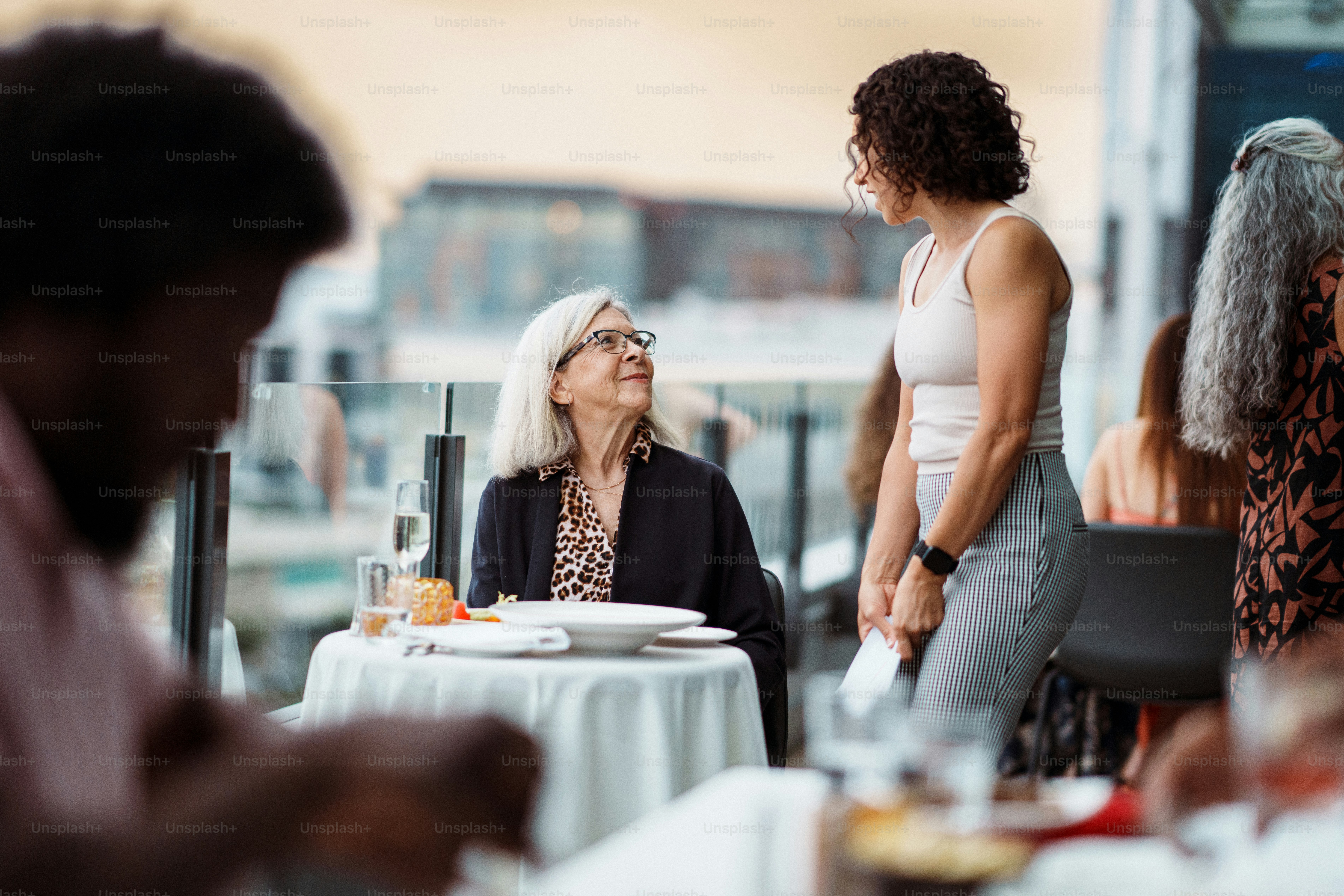 Una vibrante anciana habla con una camarera euroasiática mientras cena sola al aire libre en la terraza de la azotea de un restaurante de lujo en la ciudad al atardecer.
