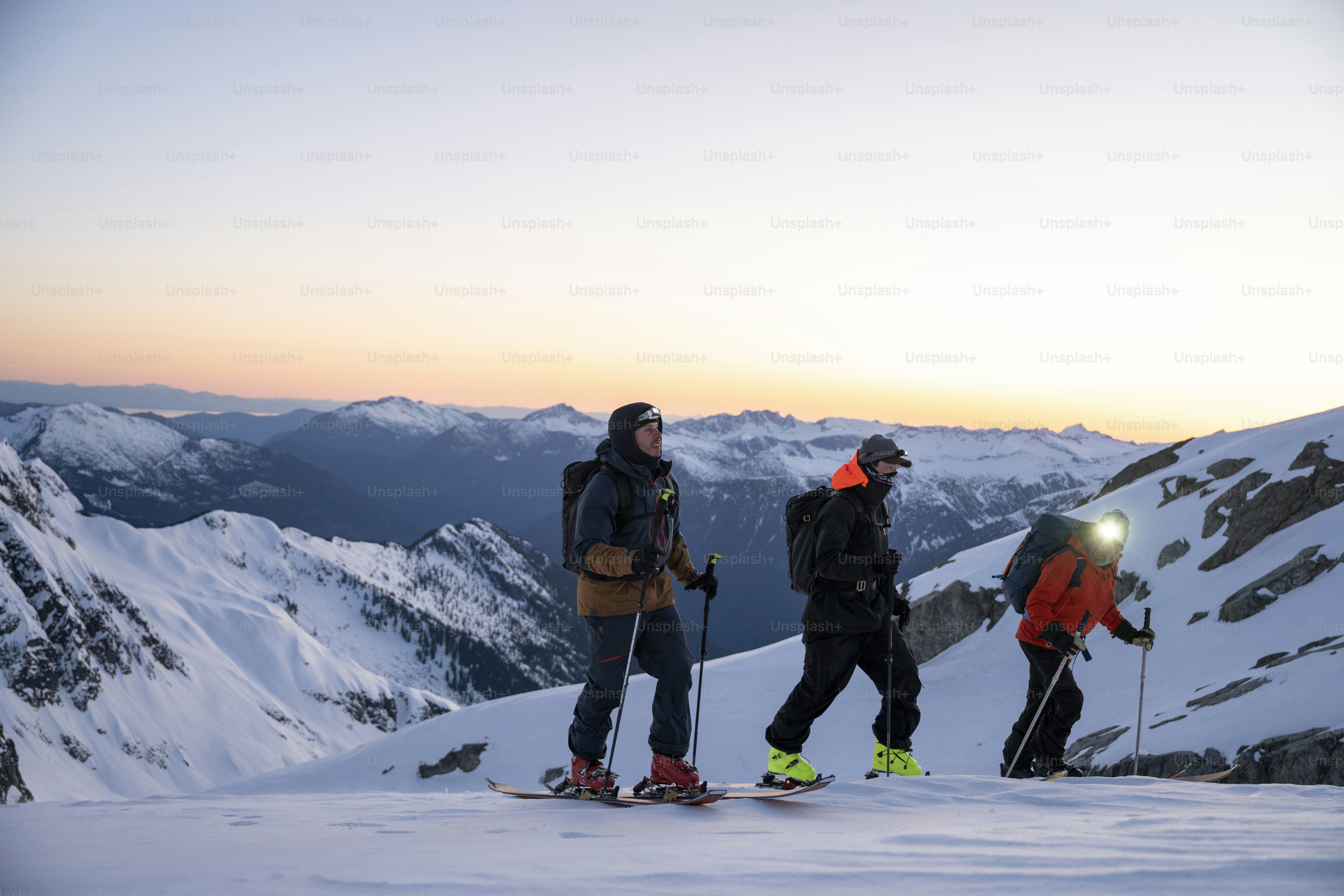 Sekelompok pemain ski pedalaman bersiap untuk menuruni gunung saat matahari terbit menggunakan lampu depan, Tantalus Range, B.C.