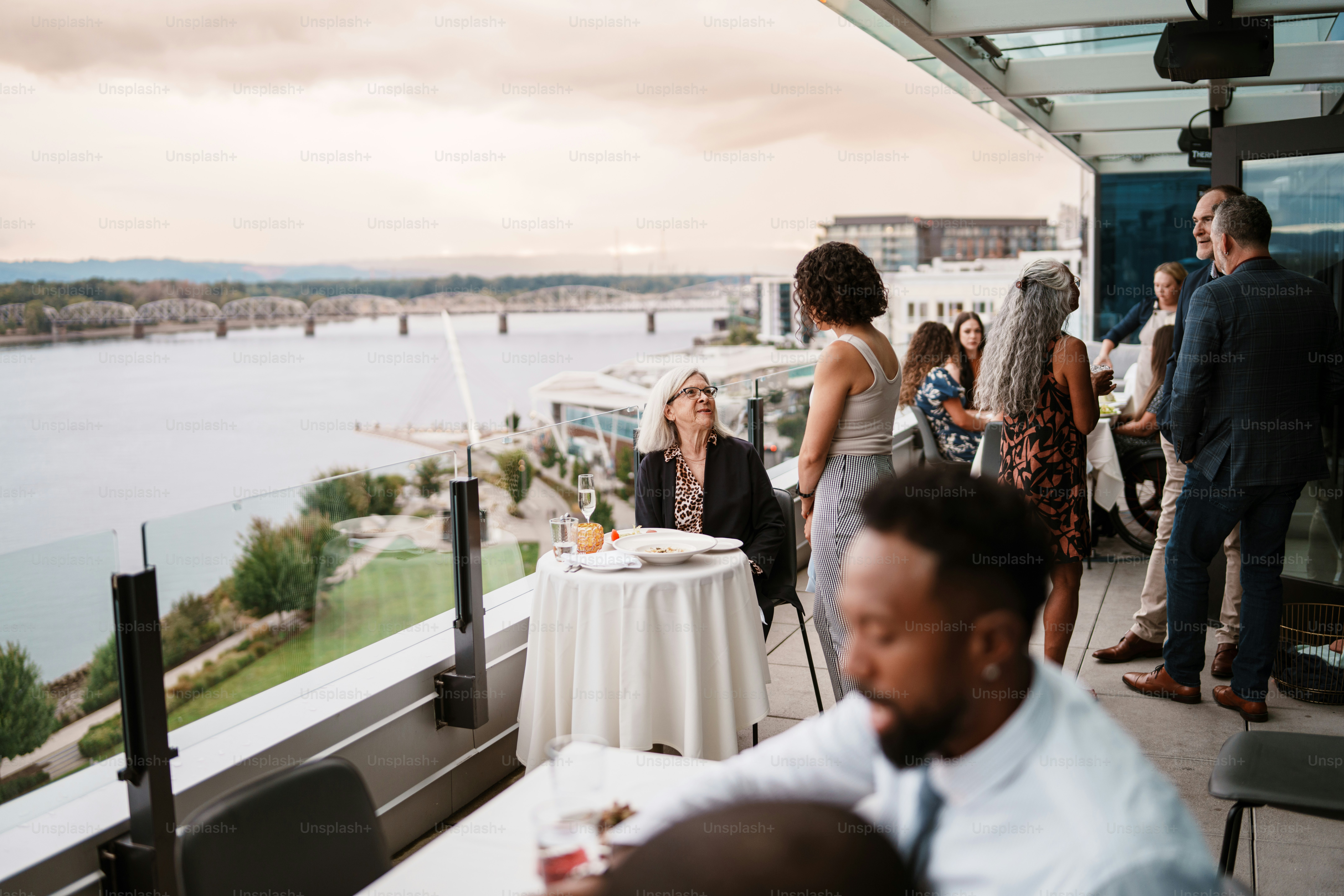 Una vibrante anciana habla con una camarera euroasiática mientras cena sola al aire libre en la concurrida terraza de la azotea de un restaurante de lujo con vistas a un río.