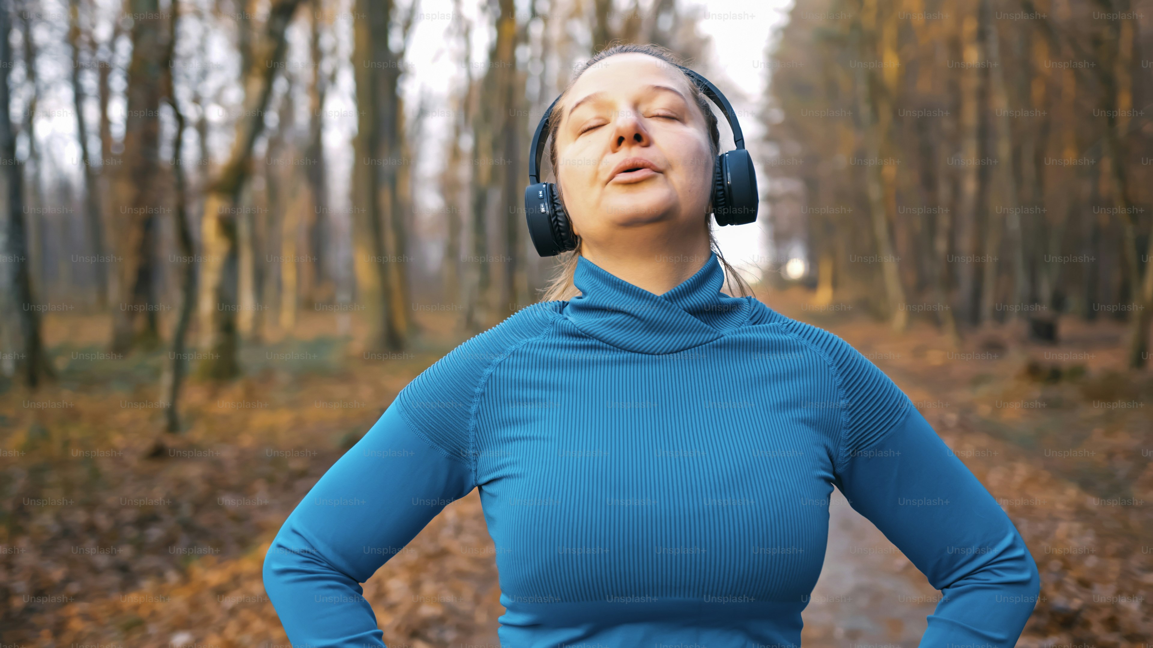 A confident Caucasian woman in her 40s wearing a blue sweater and headphones stands in a forest, mentally focusing before a jogging session on a crisp autumn day.