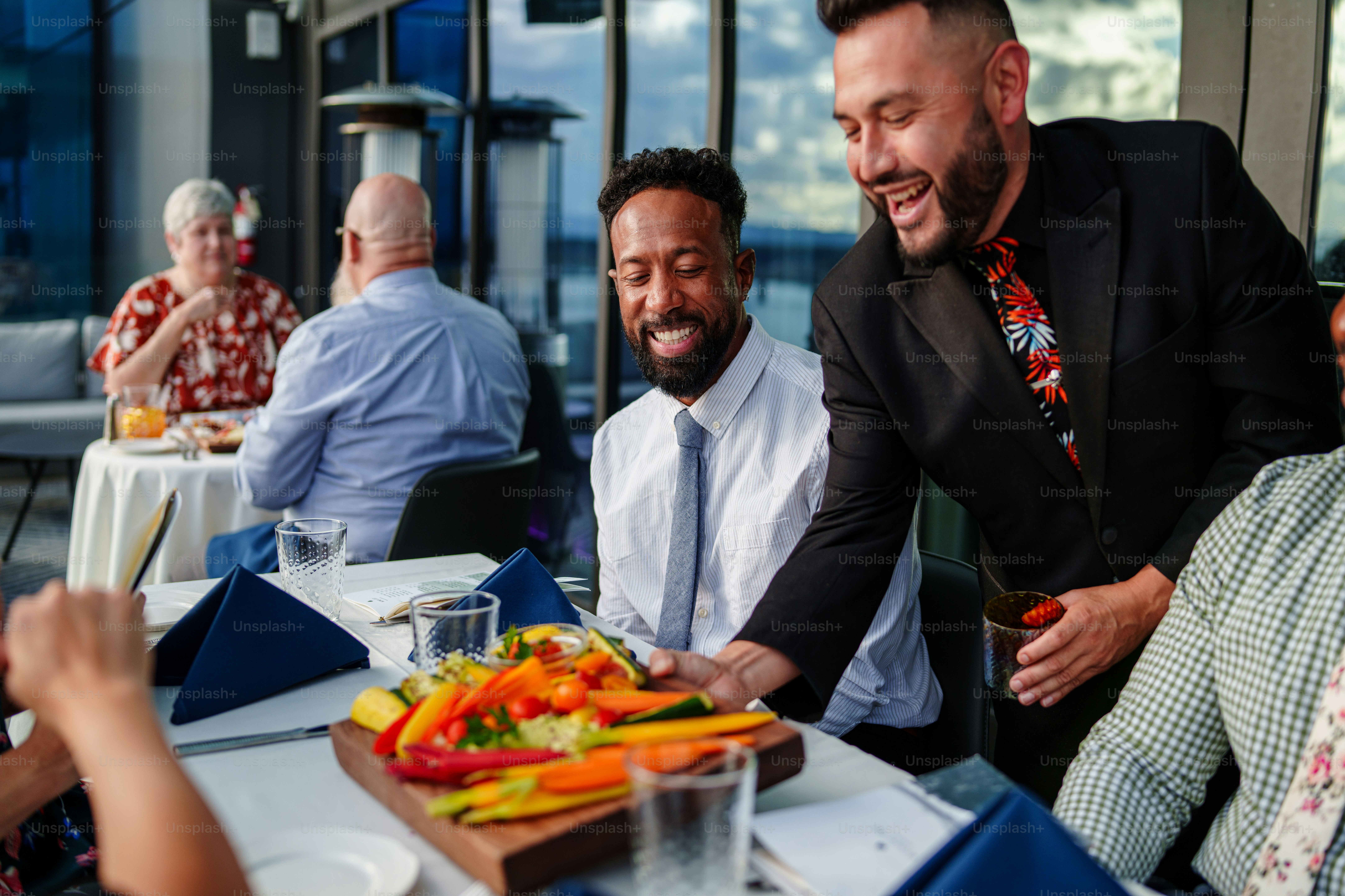 A male waiter of Hispanic descent serves a charcuterie board appetizer to a multiracial group of adults on a double date who are seated at a table on the rooftop terrace of an upscale restaurant.