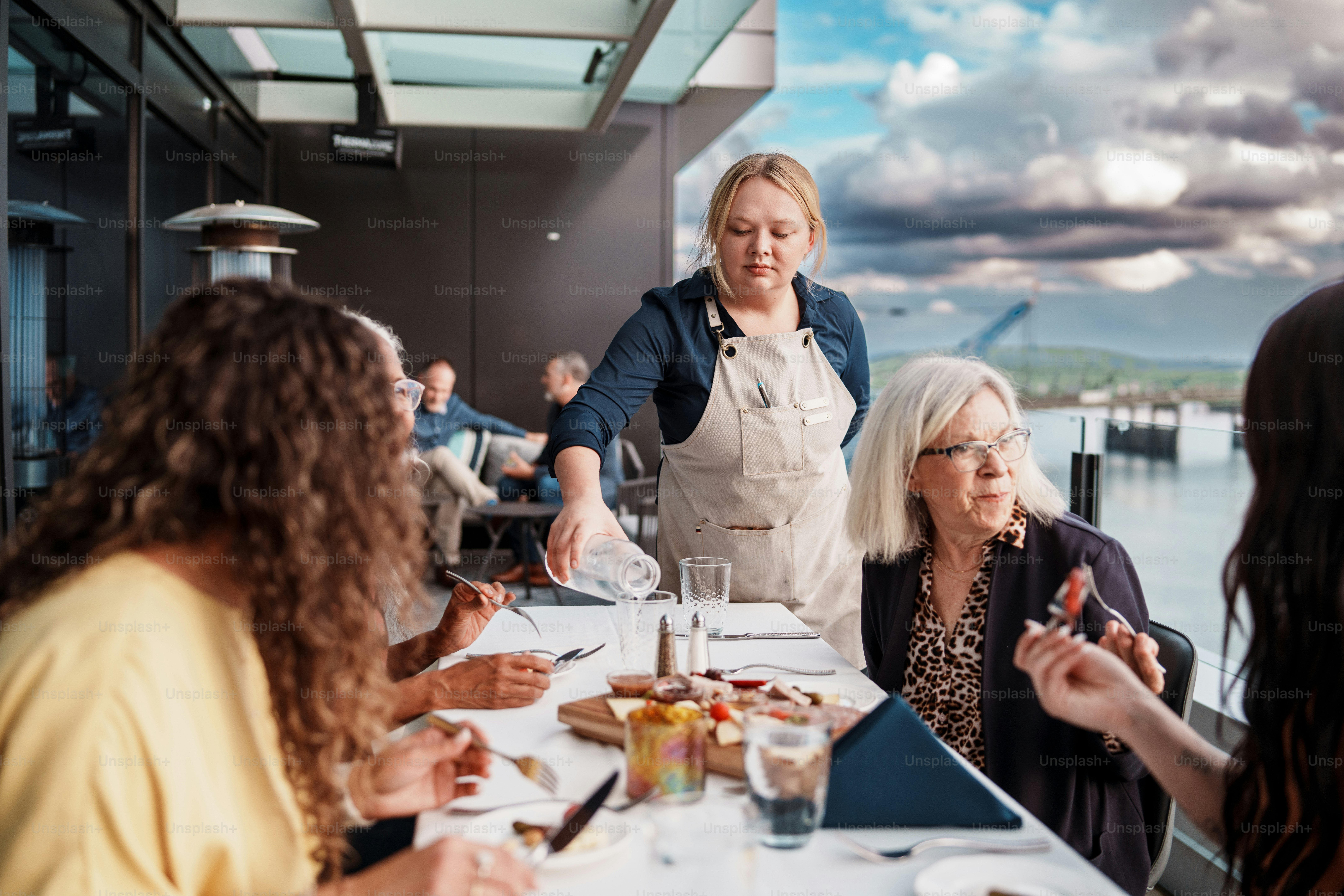 A waitress pours water into the glass of a restaurant guest seated outdoors on the rooftop patio with a group of friends.