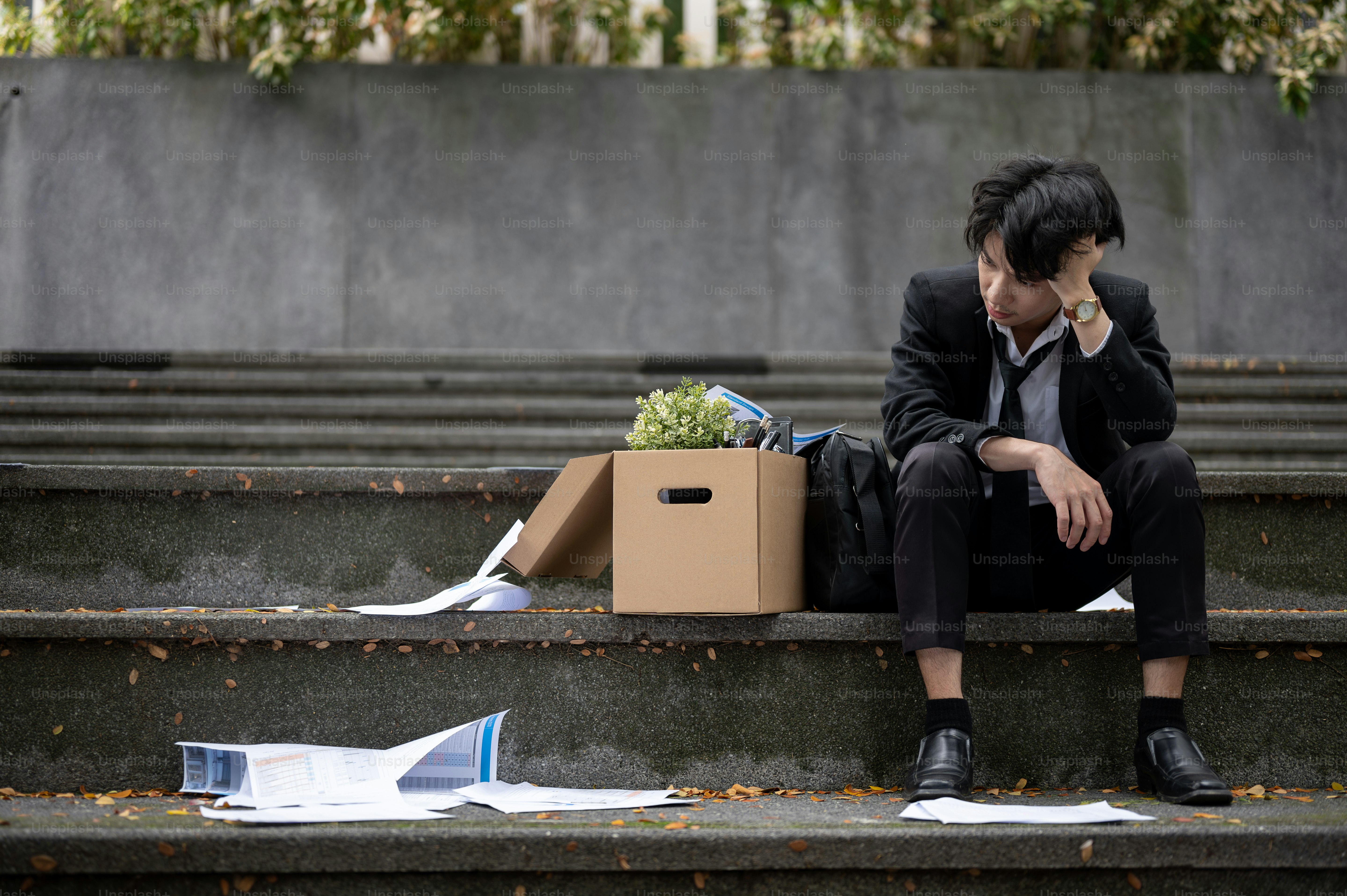 A depressed, upset, and sad young Asian unemployed businessman is sitting on the stairs in front of the corporate office with a box of his personal stuff after losing his job.