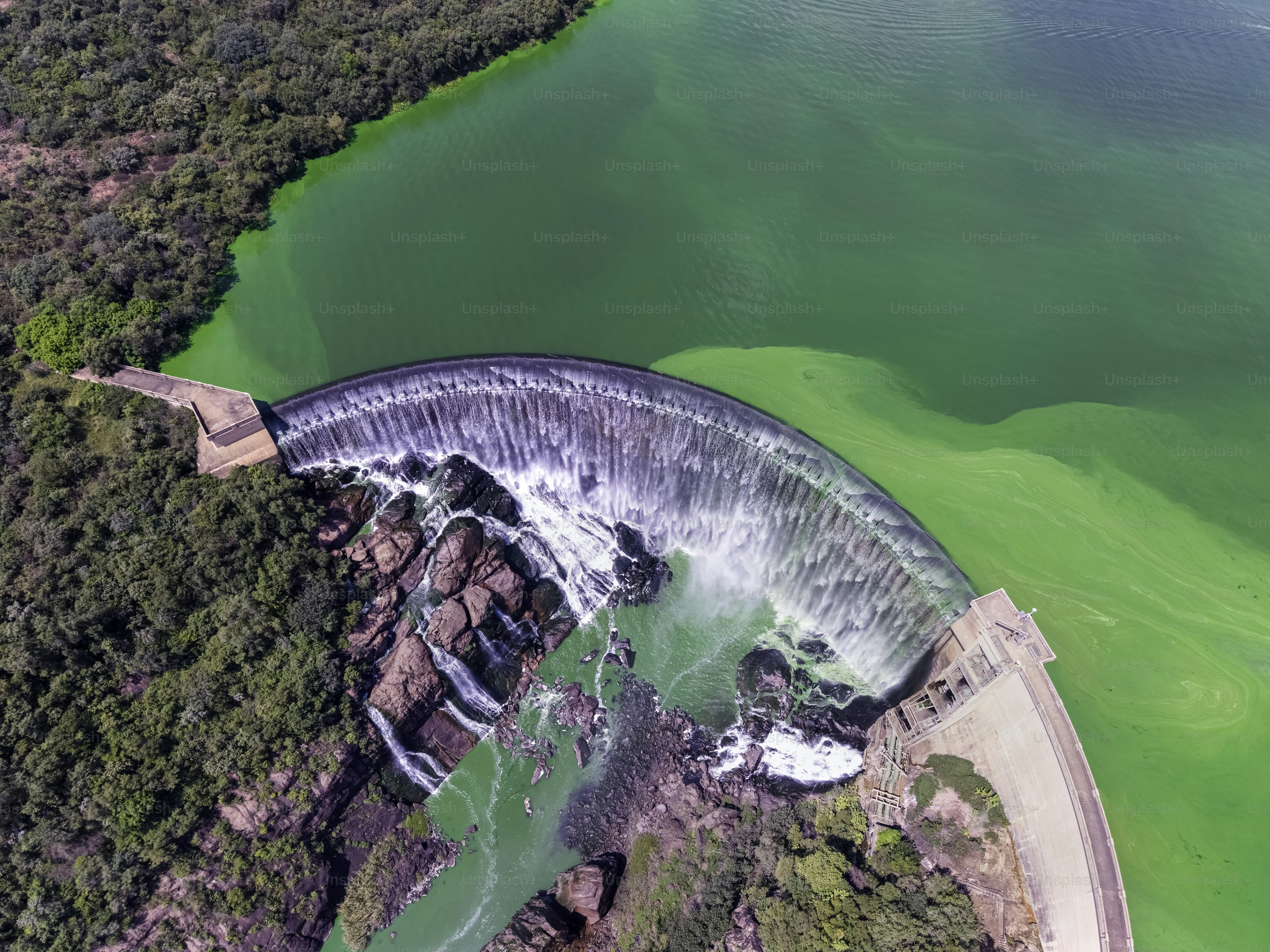 Aerial view of a curved dam spilling water into a rocky green river