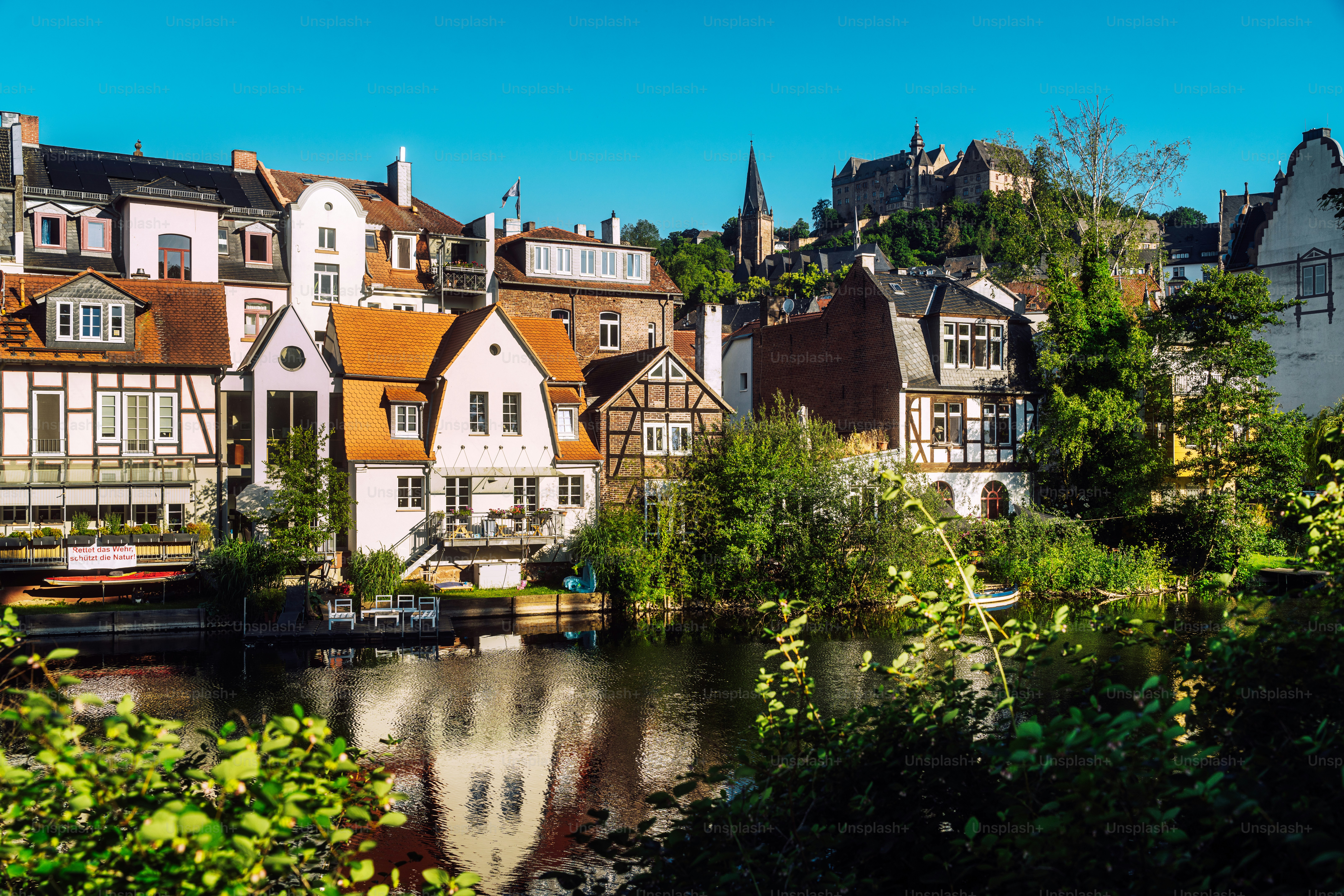Blauer Sommermorgen: Historisches Marburg mit Blick auf die Lahn mit Wehr im Vordergrund und Schloss im Hintergrund.