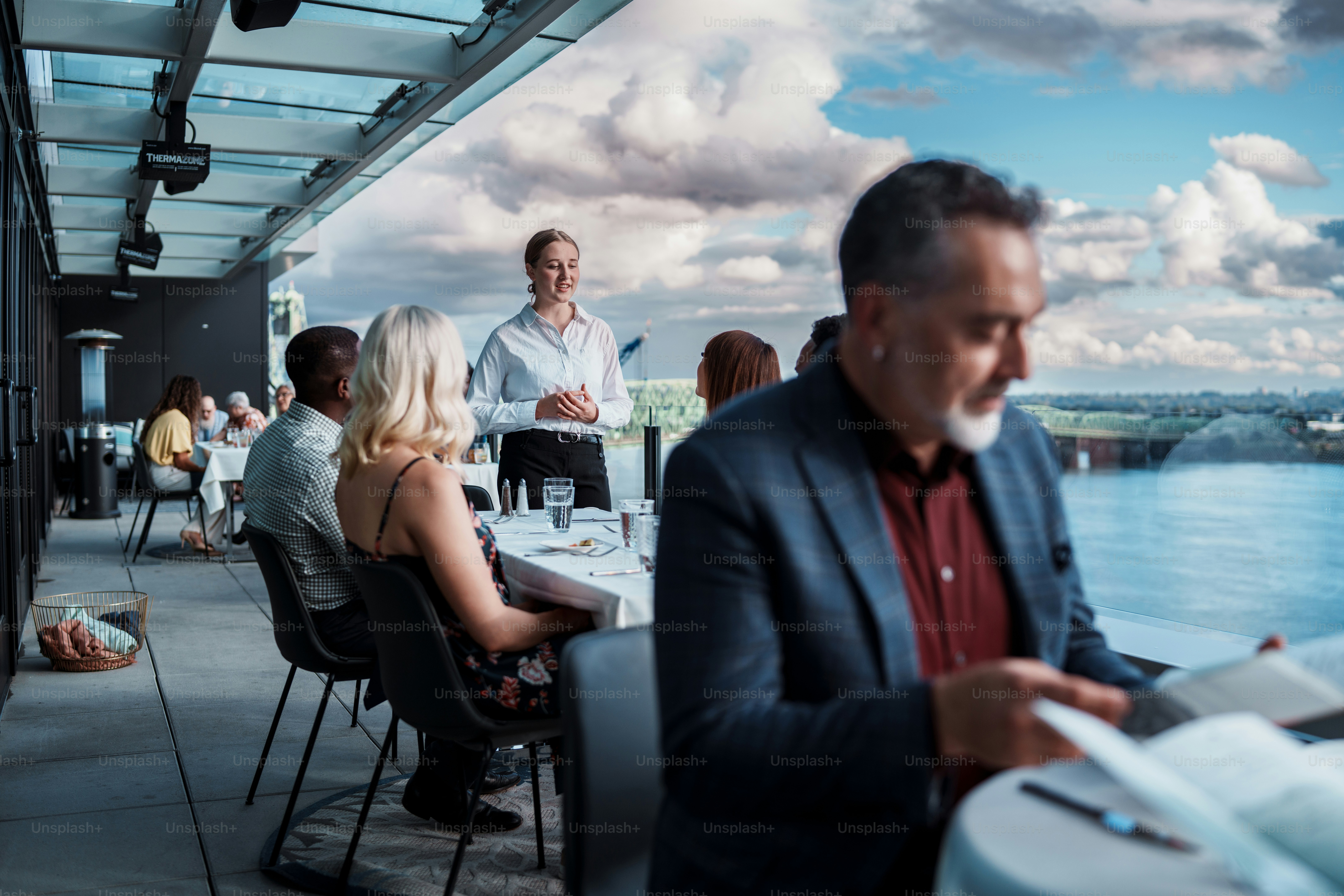 La anfitriona de un restaurante conversa con un grupo multirracial de amigos que cenan juntos en la terraza de la azotea de un restaurante de lujo en una noche nublada pero cálida.
