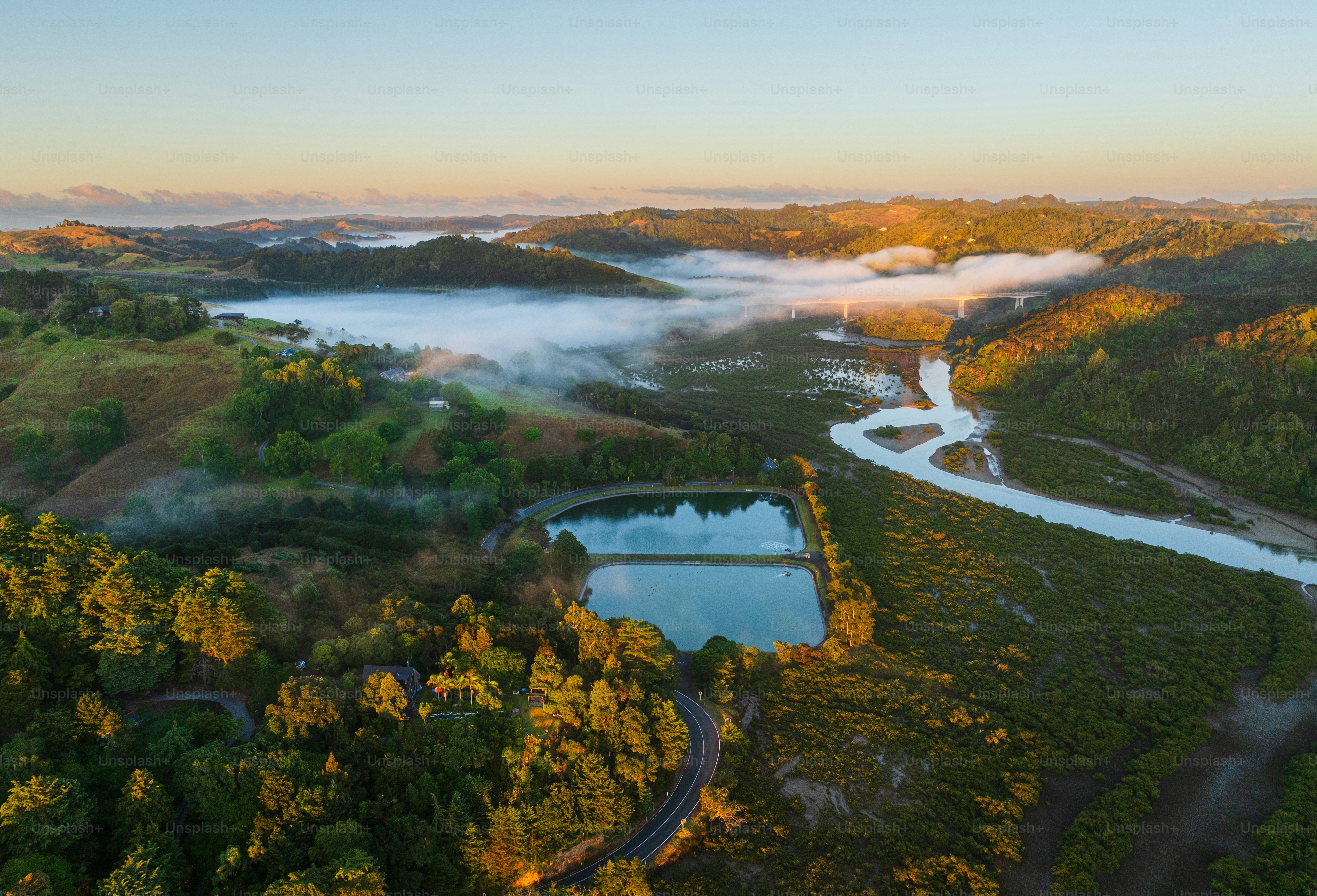 Aerial view of a scenic landscape with mist-covered hills, meandering river, and a bridge under a clear sky at sunrise in Waiwera, Auckland, New Zealand.