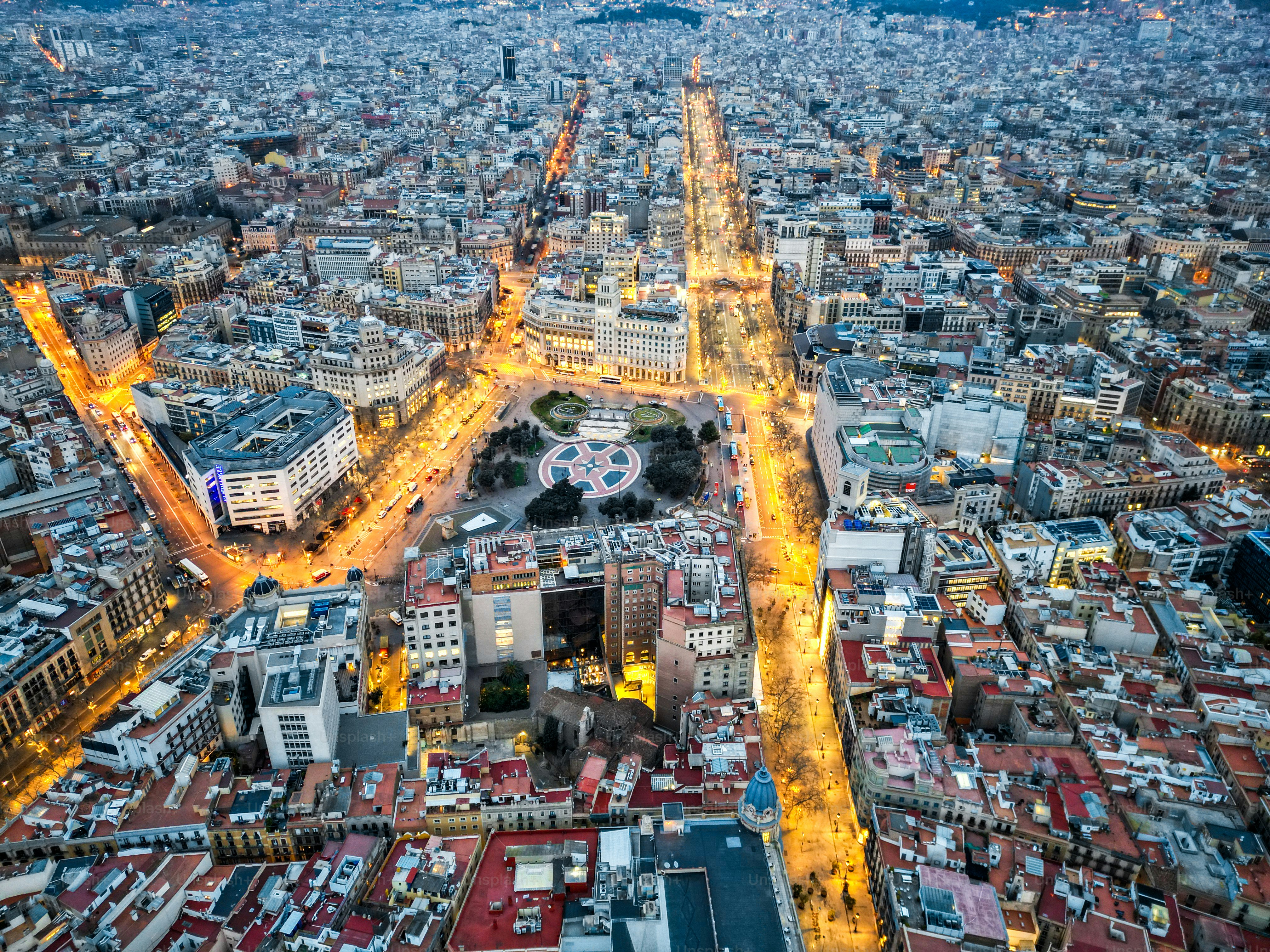 The cityscape of Barcelona unfolds in a blend of vibrant street lights and dense architecture, showcasing an aerial perspective of urban life as evening sets in.