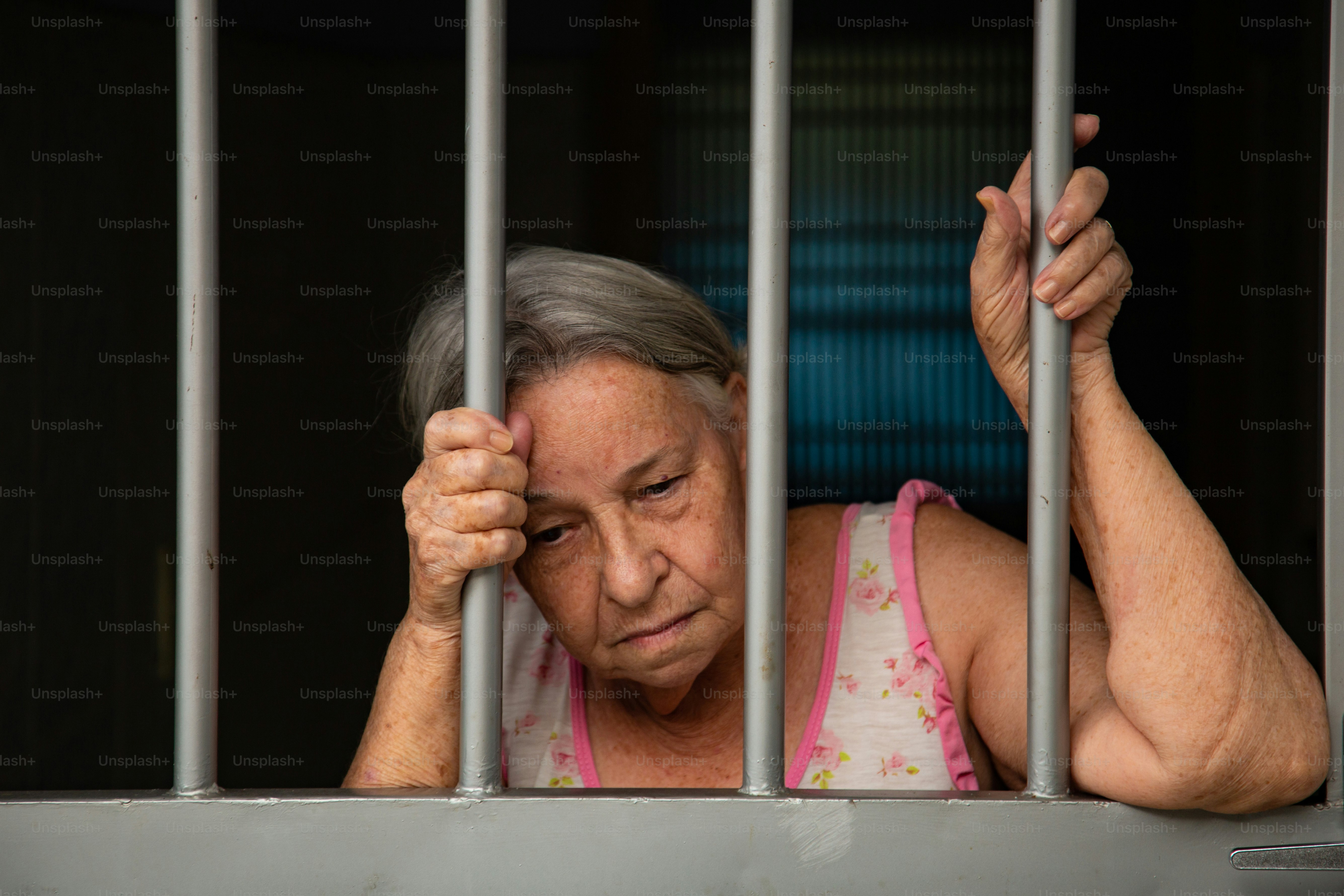 Goiânia, Goias, Brazil – March 05, 2022:  Elderly woman with sad face behind bars at the gate.