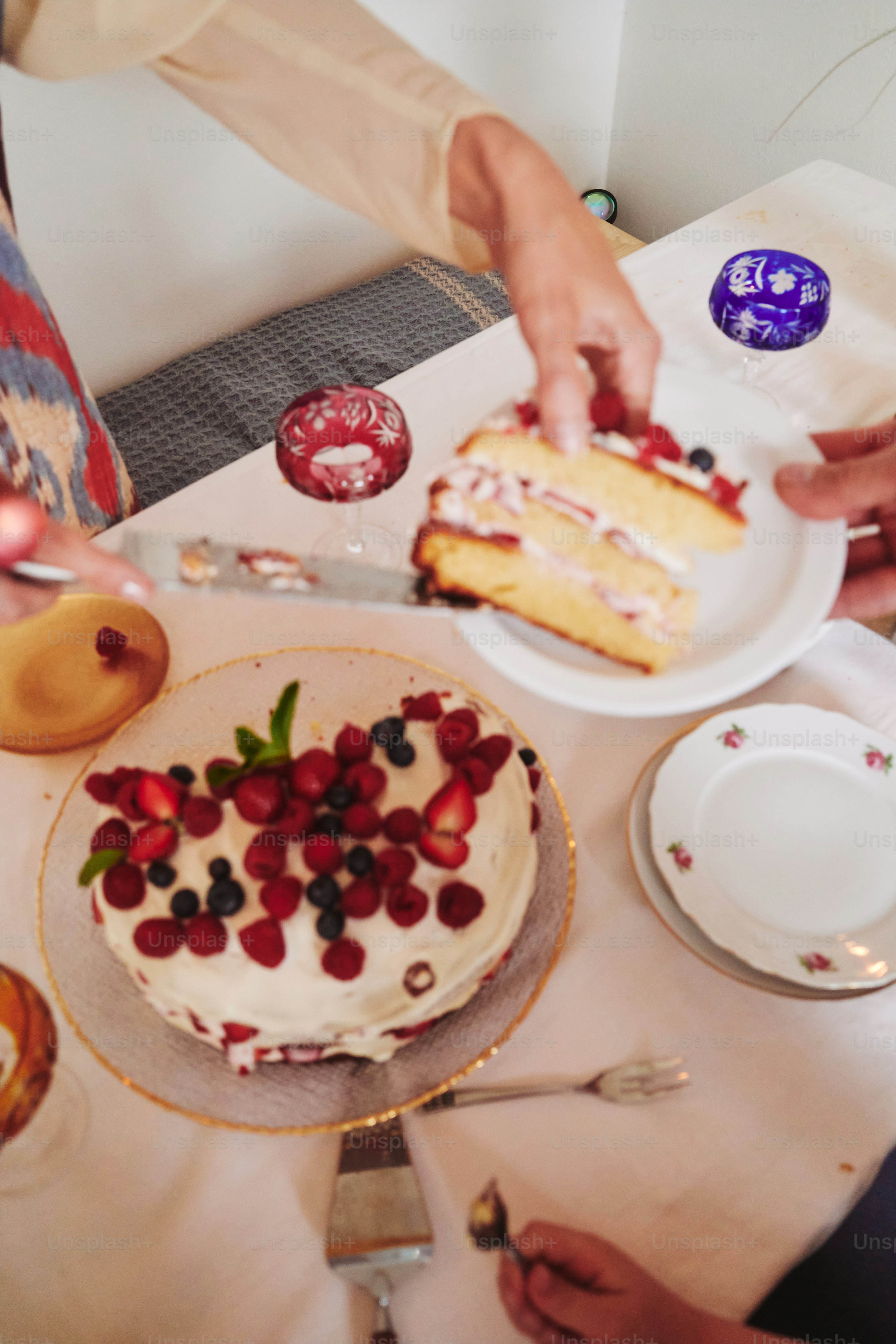 Hands serving a berry-topped cake at a table.