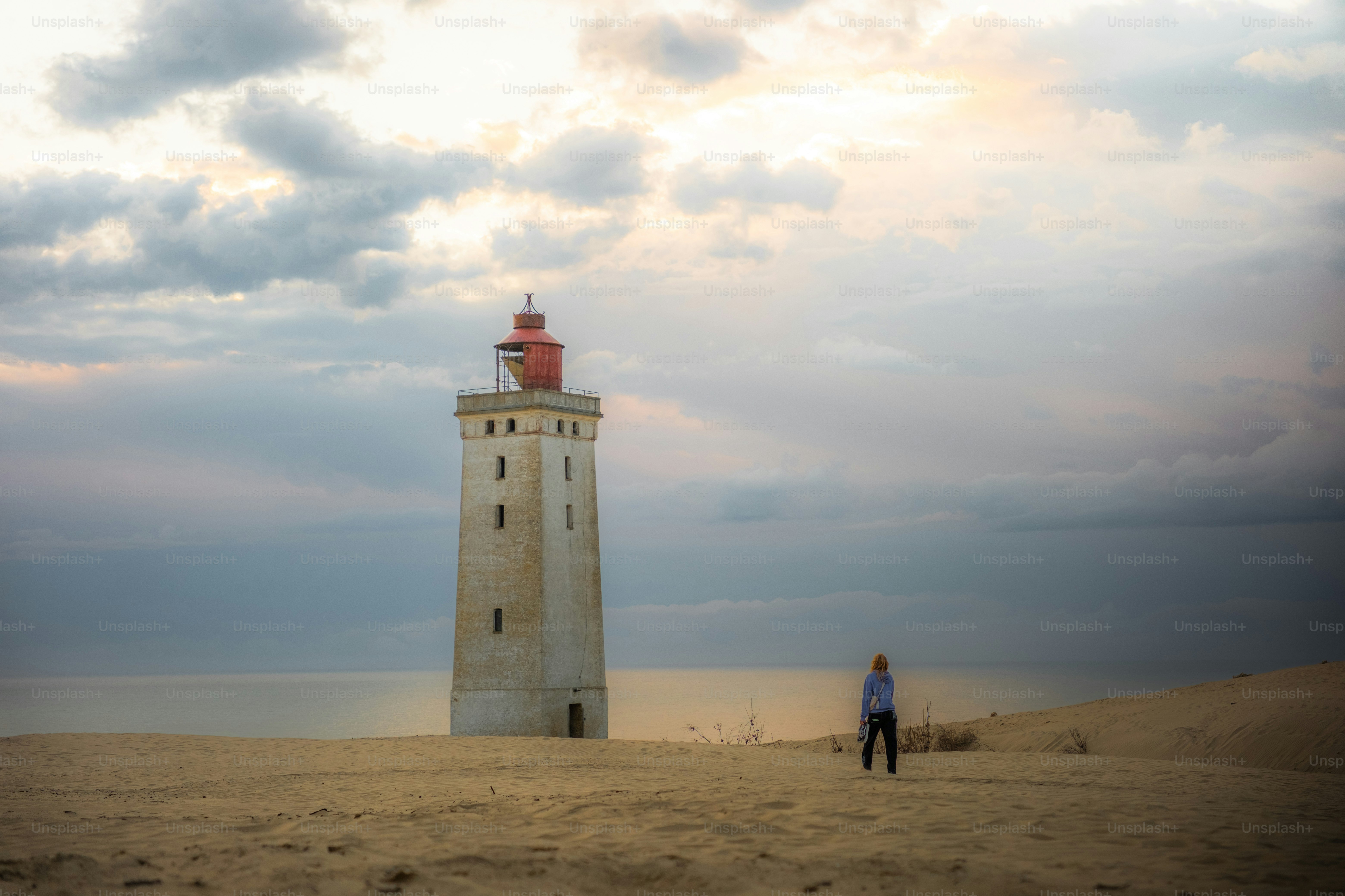 Person standing by lighthouse on sandy beach at sunset