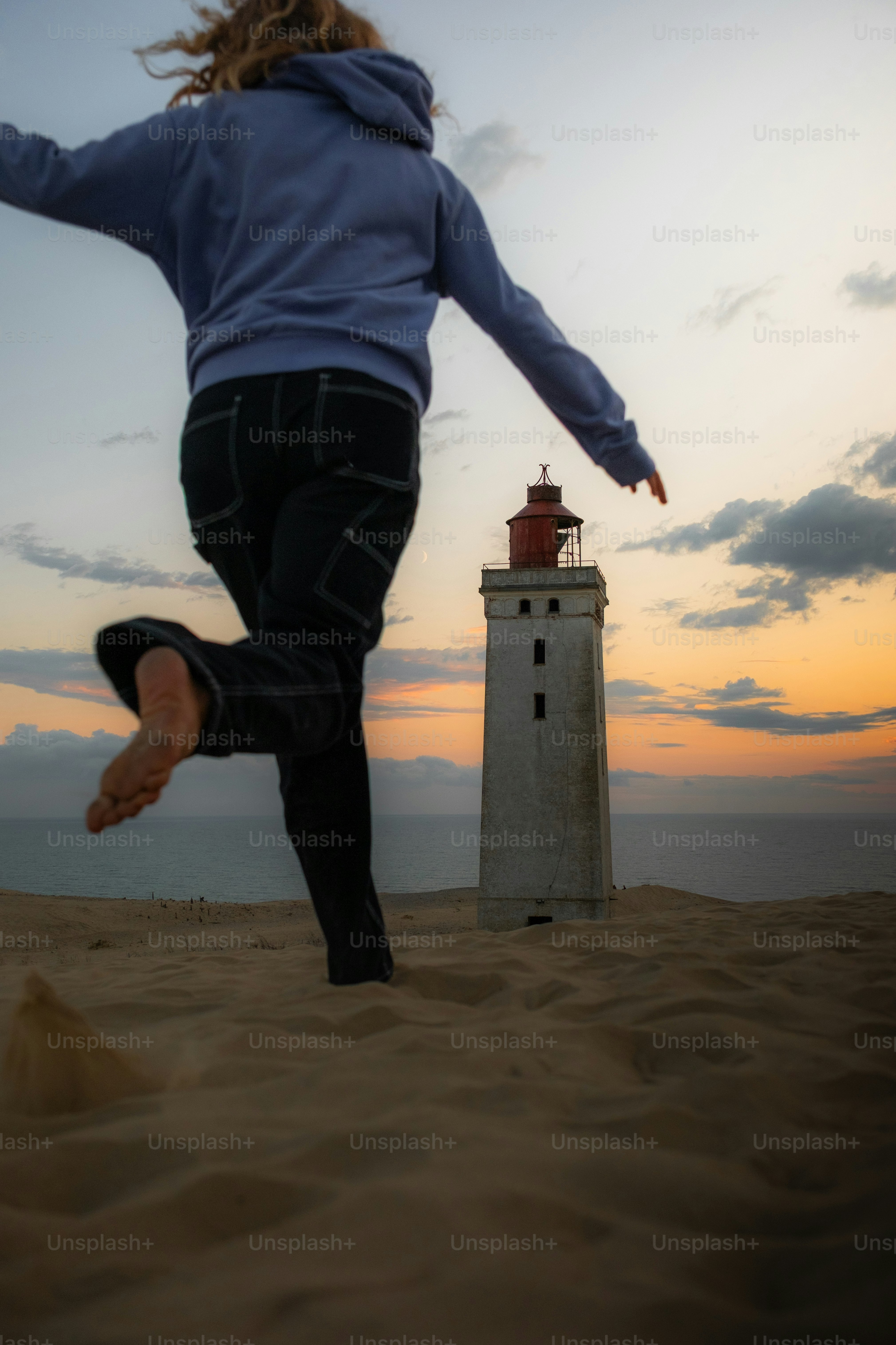 Person jumping on sand dune near lighthouse at sunset