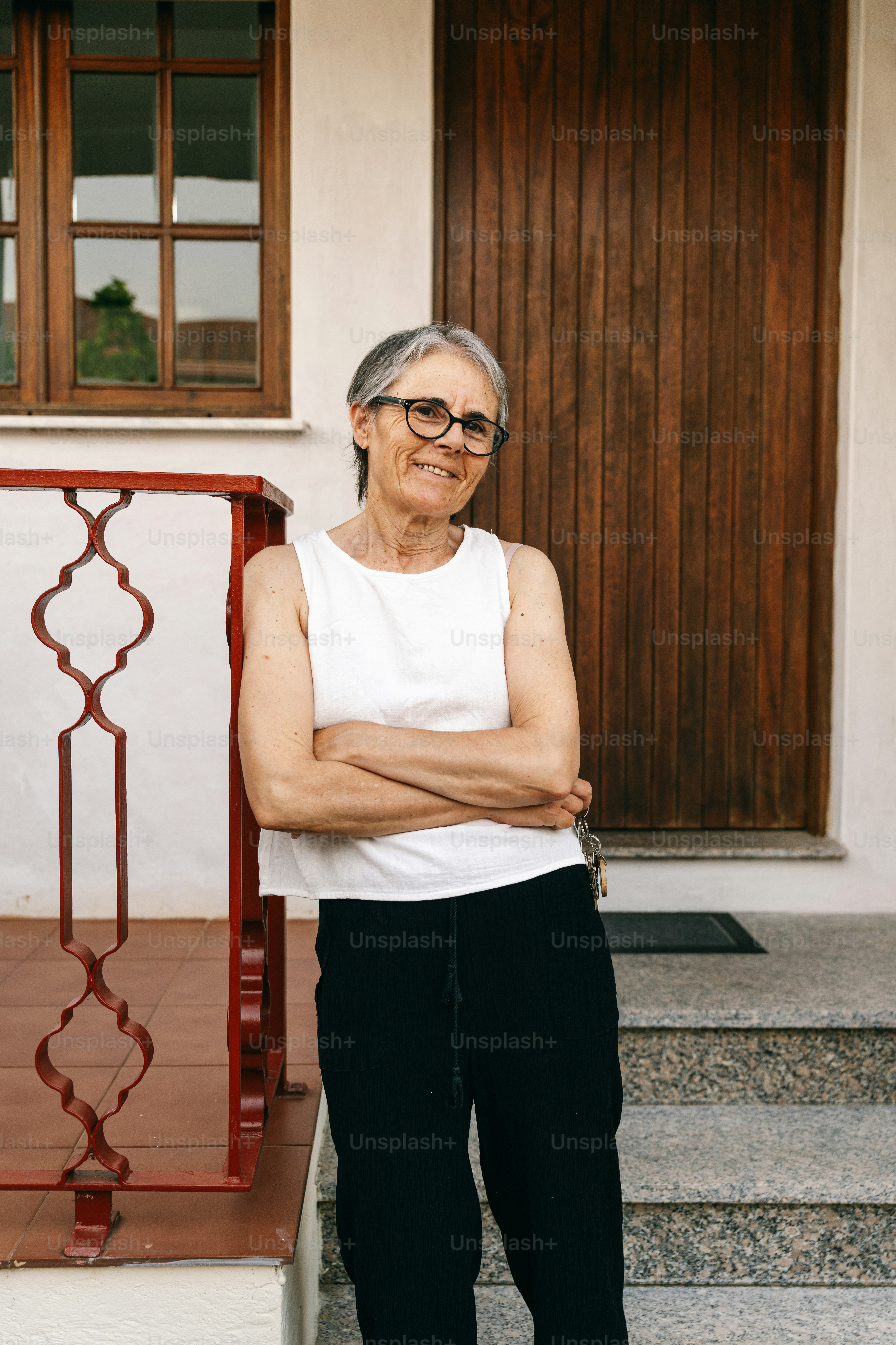 Elderly woman with glasses stands with arms crossed.