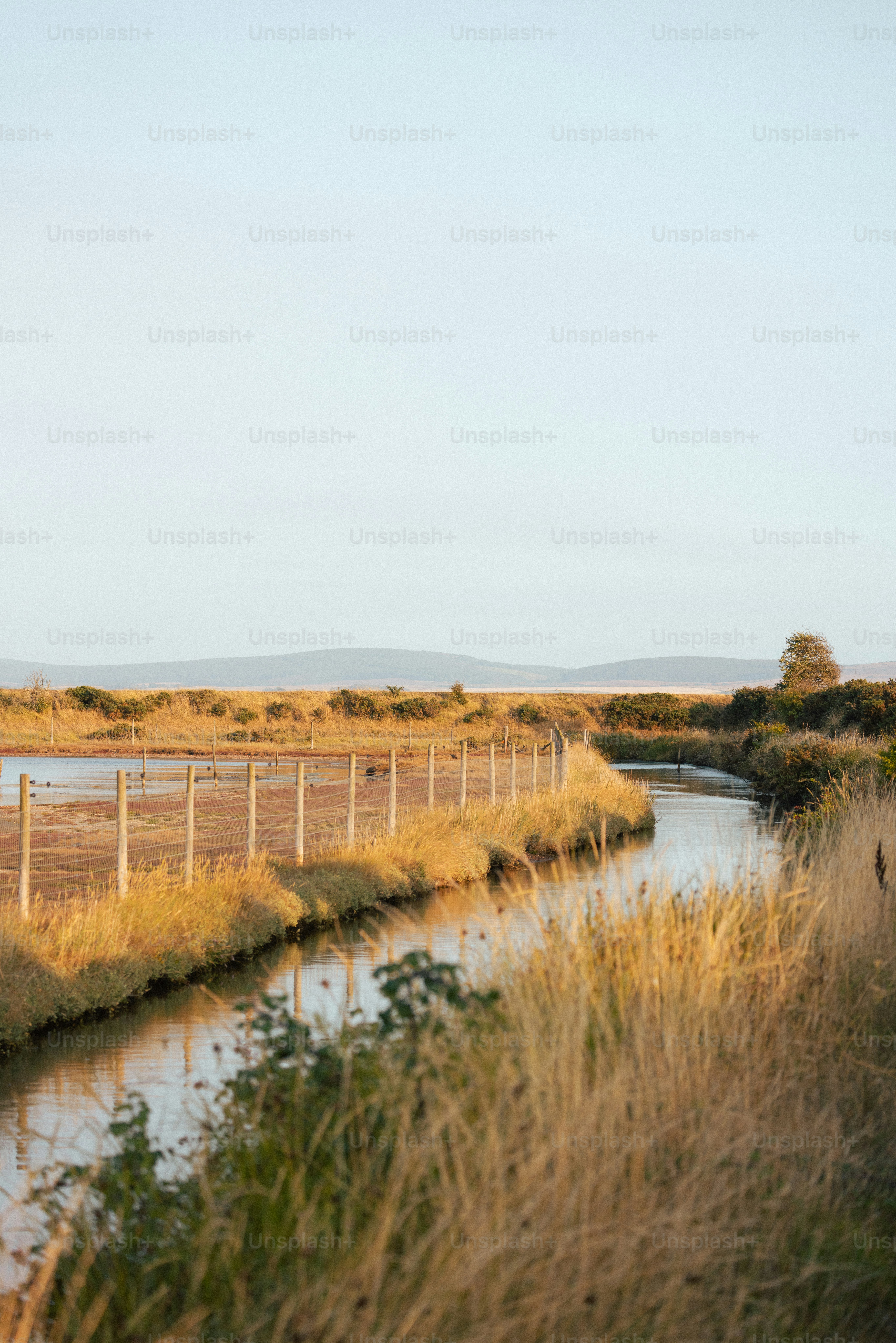 A calm river flows through a grassy field with a fence.