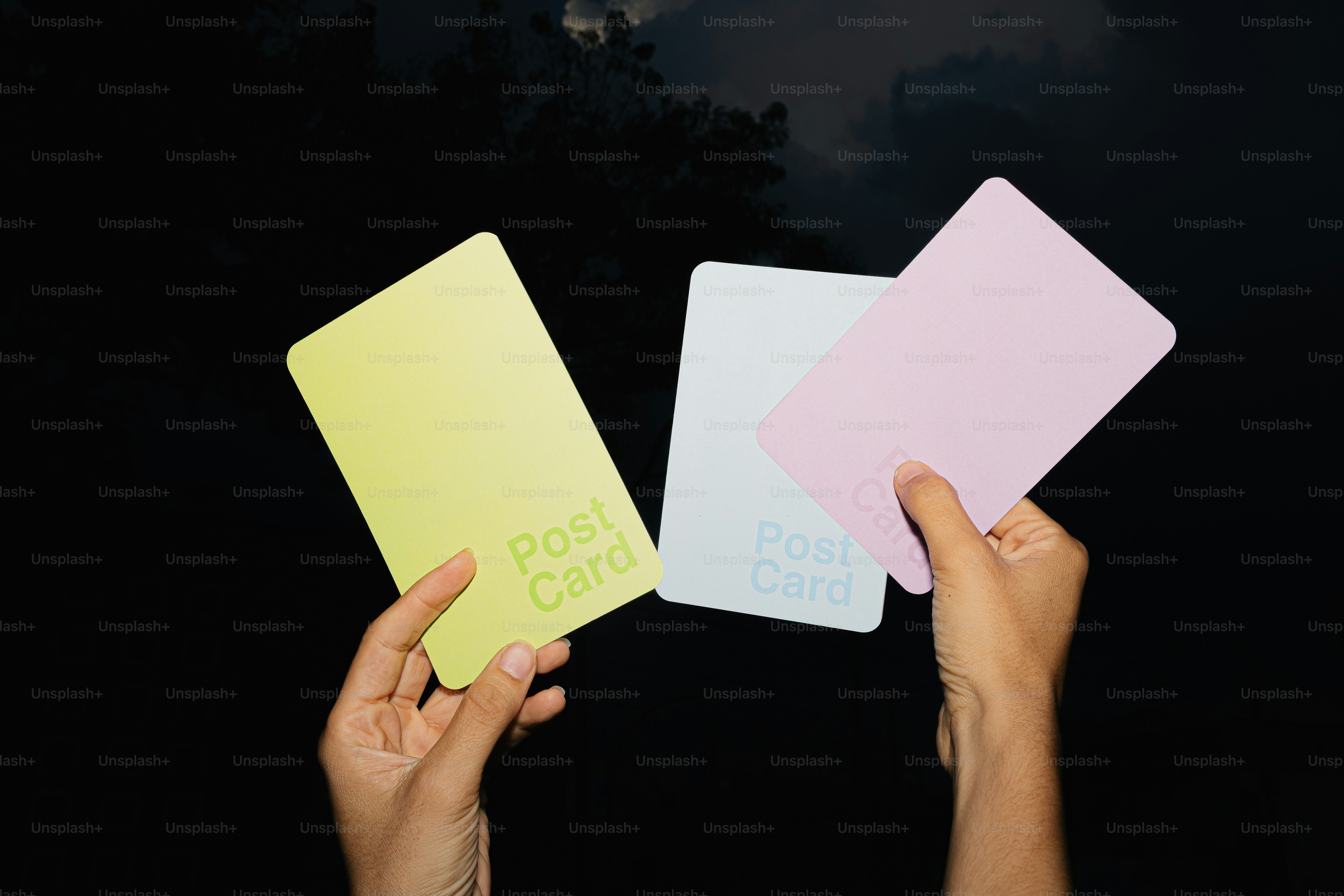 Hands hold three blank pastel postcards against dark background.