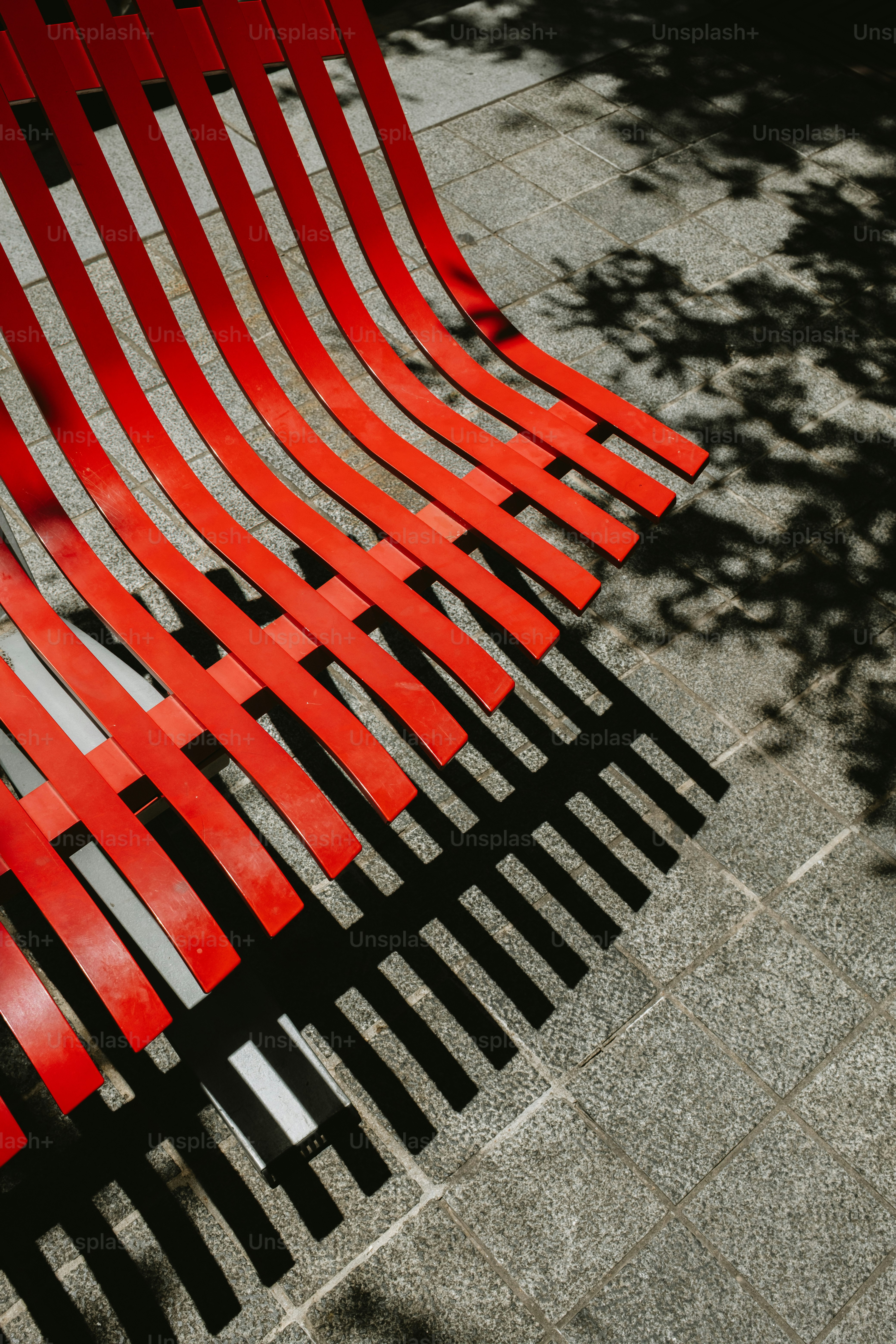 Banco de ripas vermelhas com luz solar e sombras manchadas.