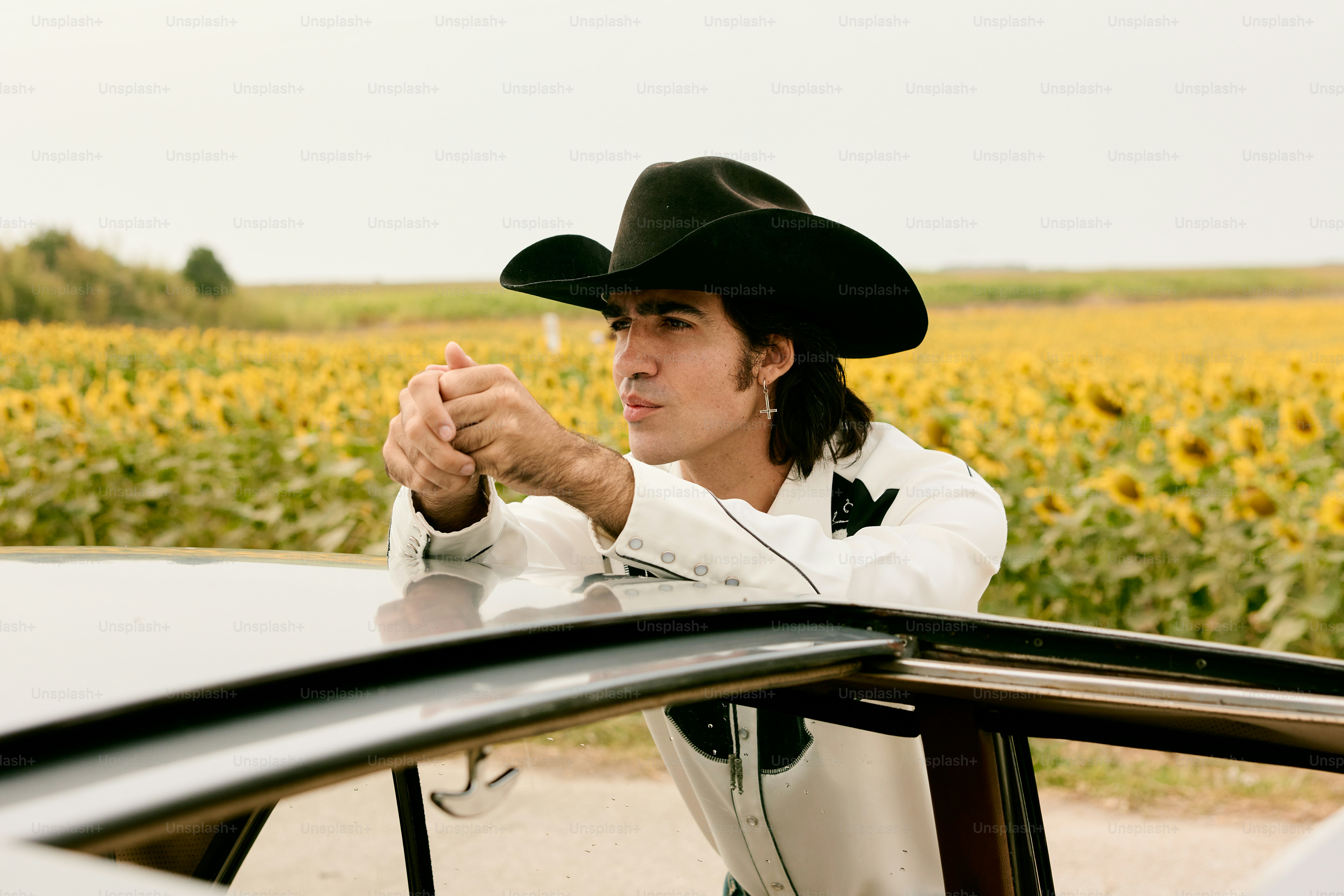 카우보이 모자를 쓴 남자가 해바라기를 뒤에 두고 차에 기대어 있습니다.