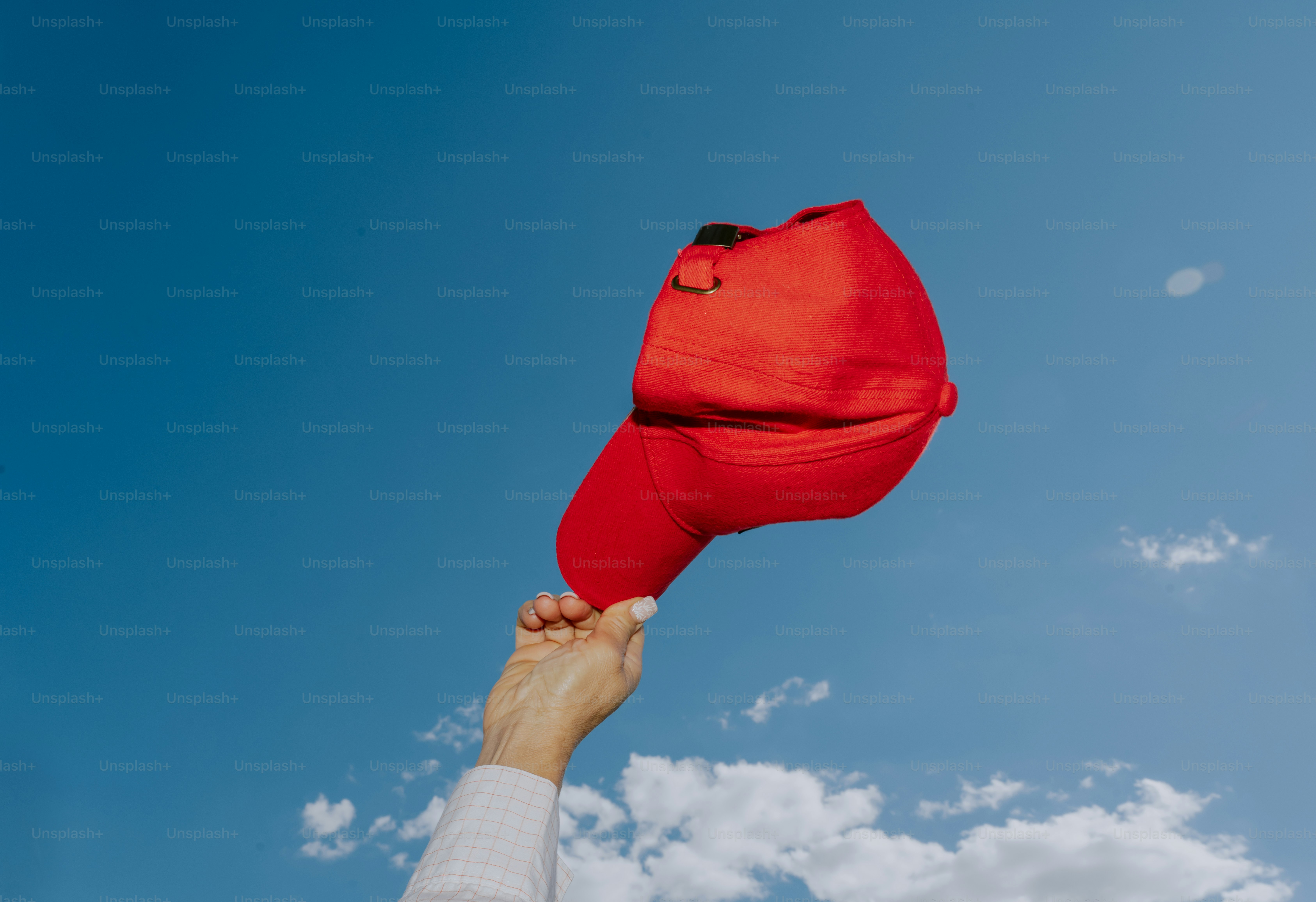 Hand holding a red baseball cap against blue sky.