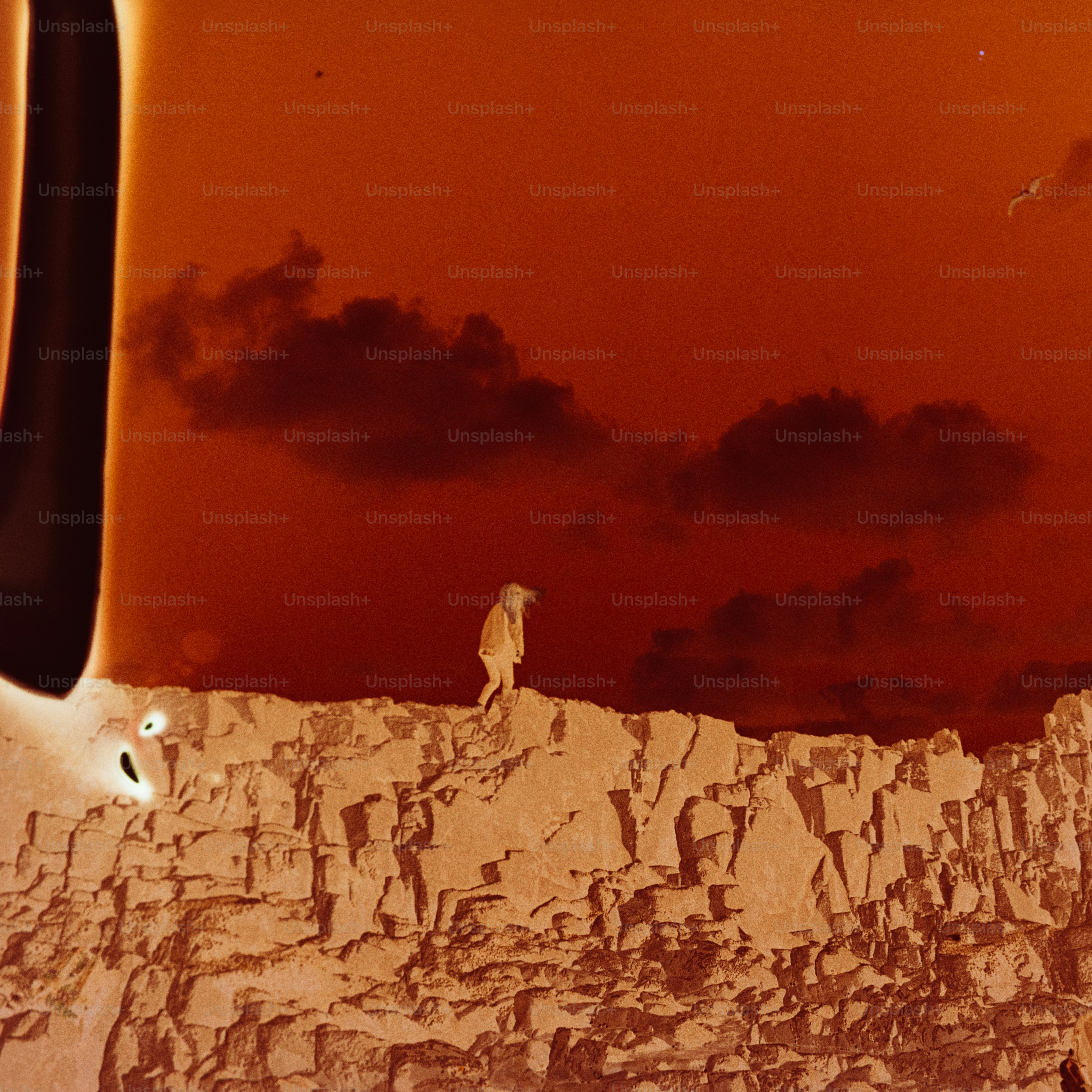 Person standing on rocky cliff under orange sky.