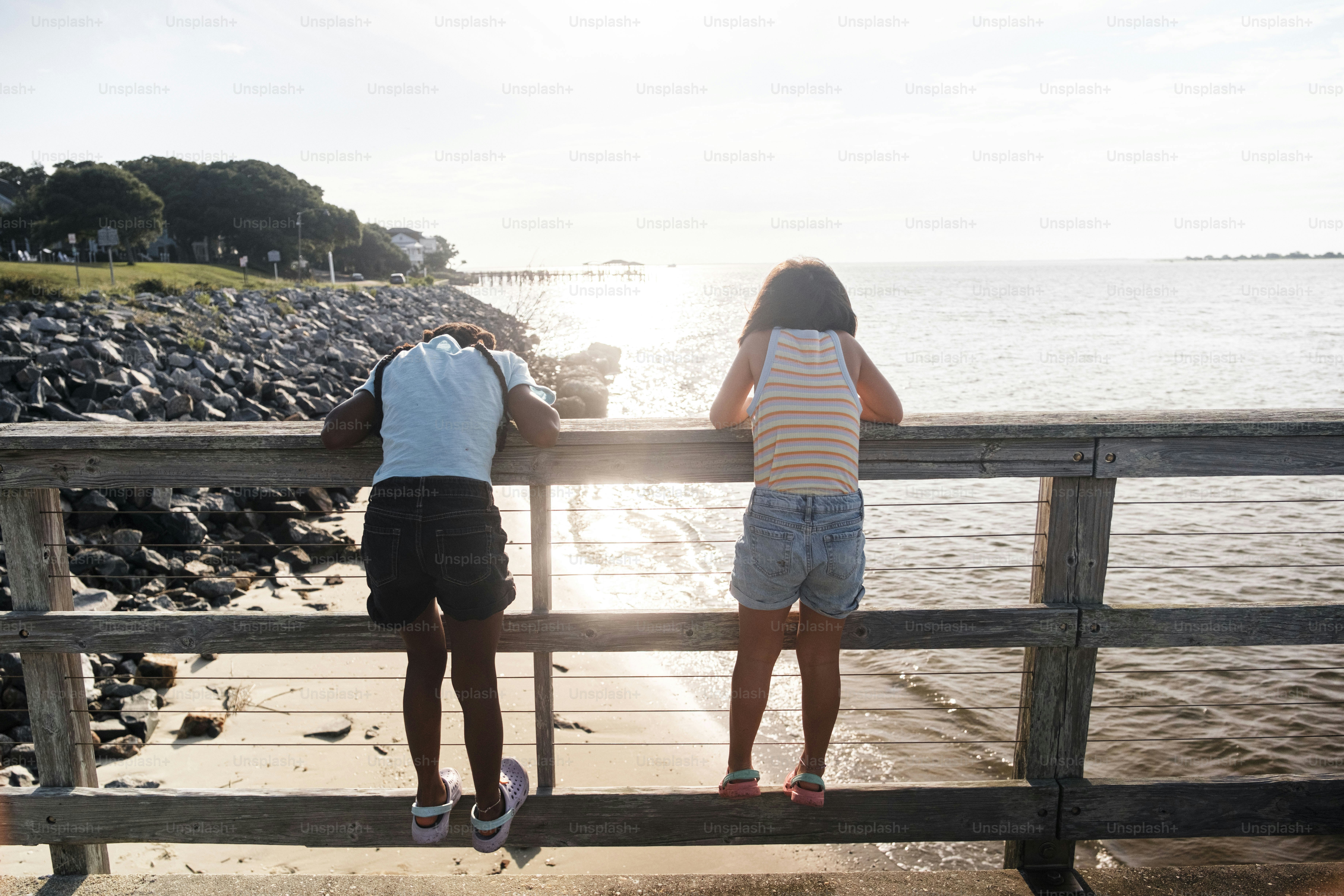 Two children lean over a wooden railing by the sea.