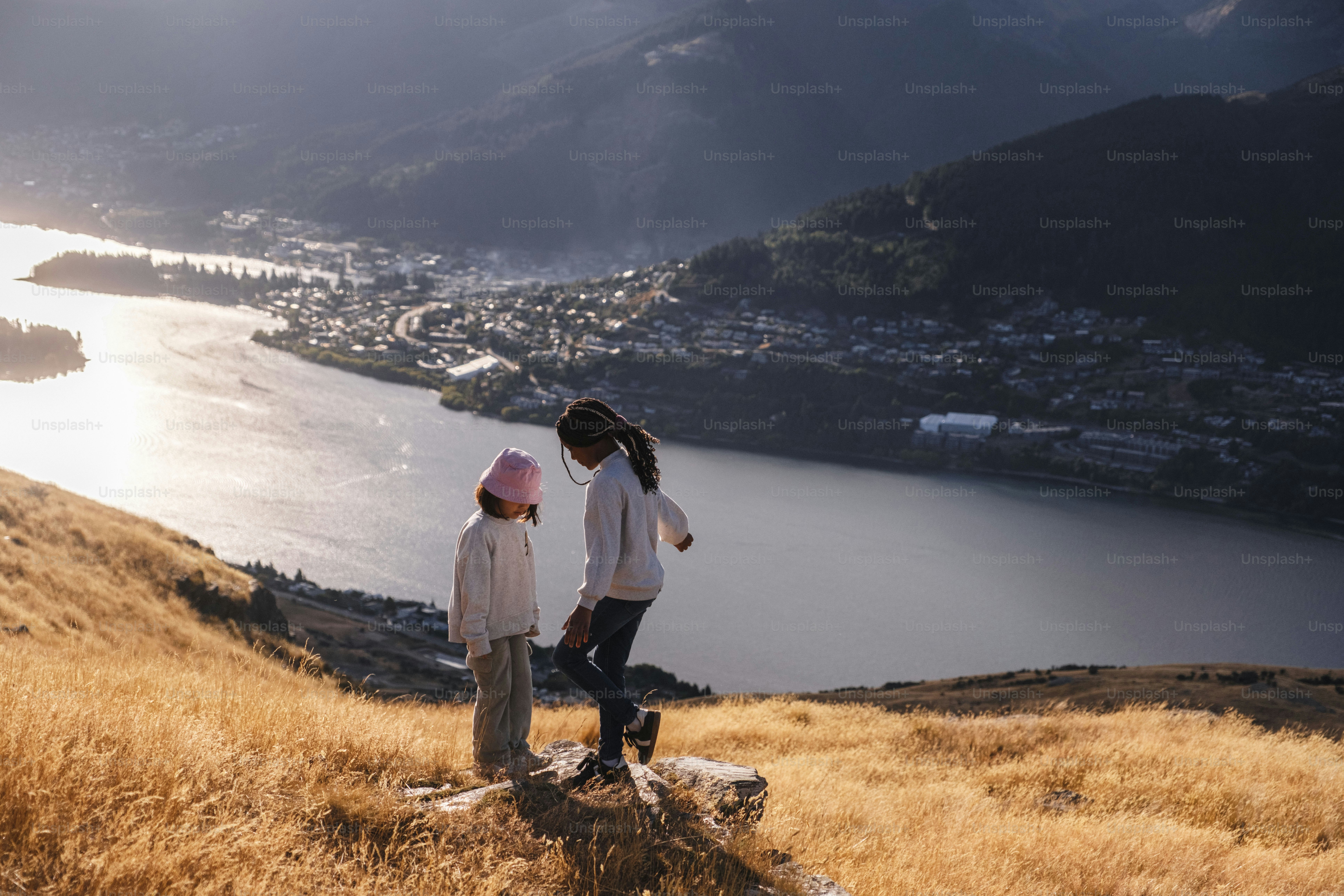 Two children stand on a grassy hill overlooking a lake.