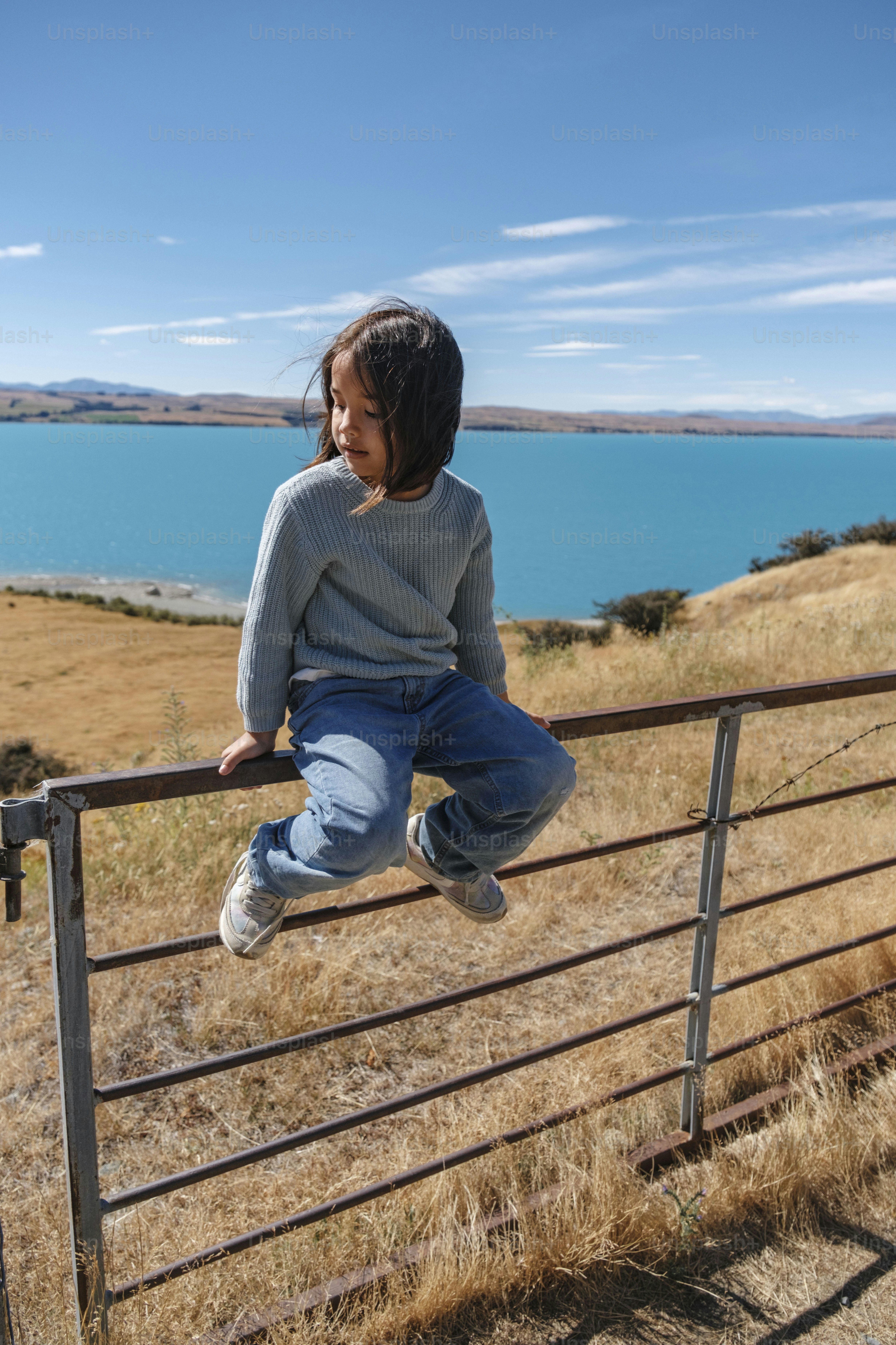 Young girl sits on a metal fence overlooking lake.