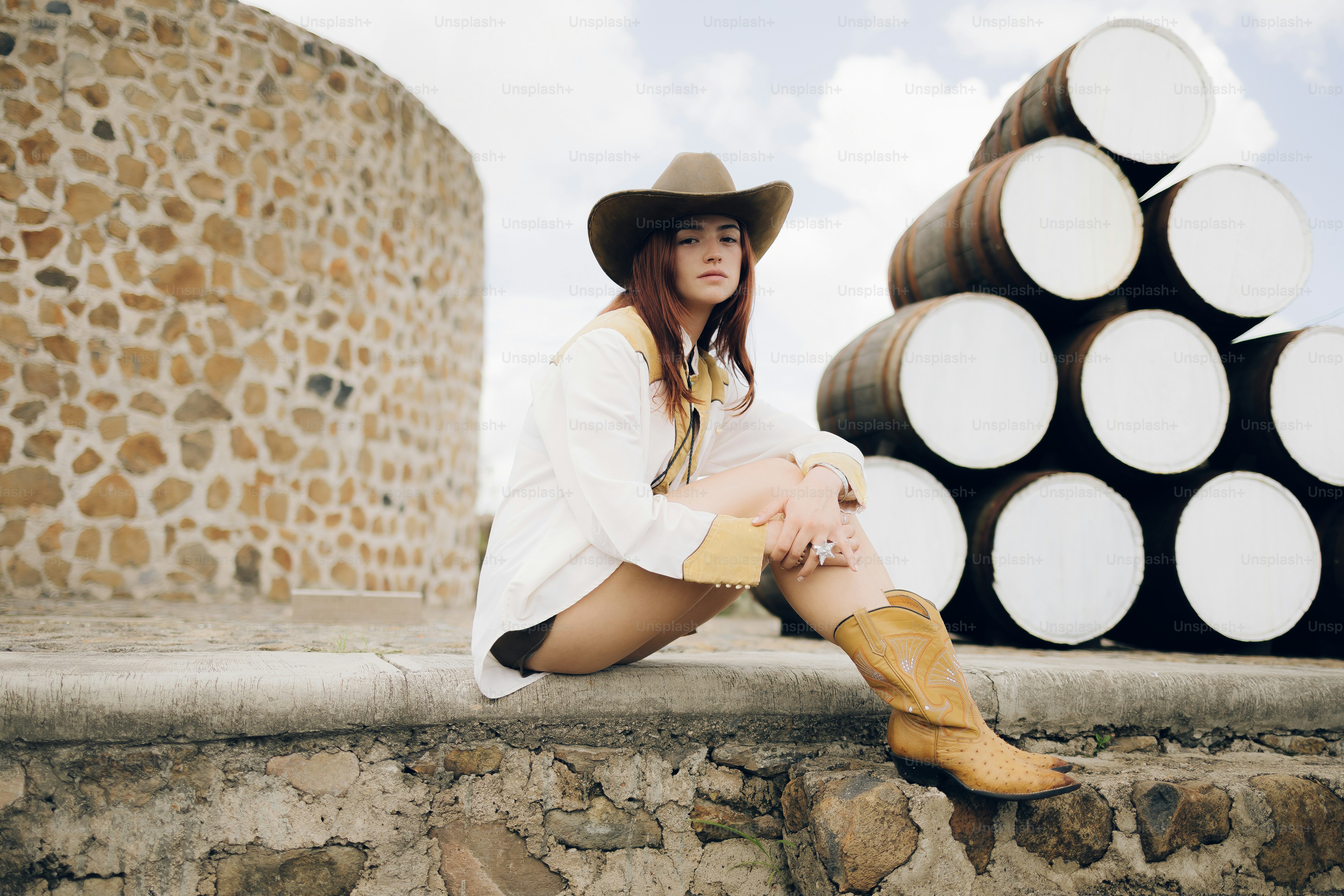 Woman in cowboy hat and boots sits by barrels.