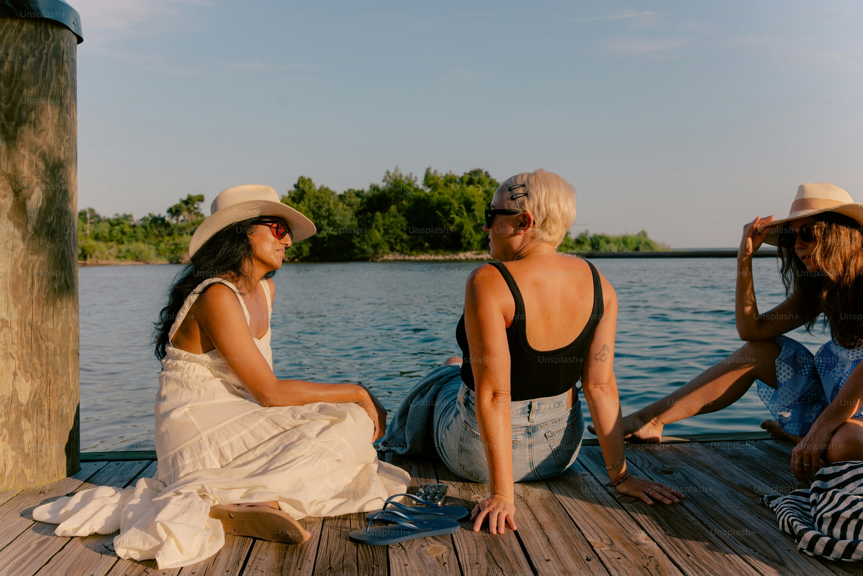 Three women sit on a wooden dock by the water.