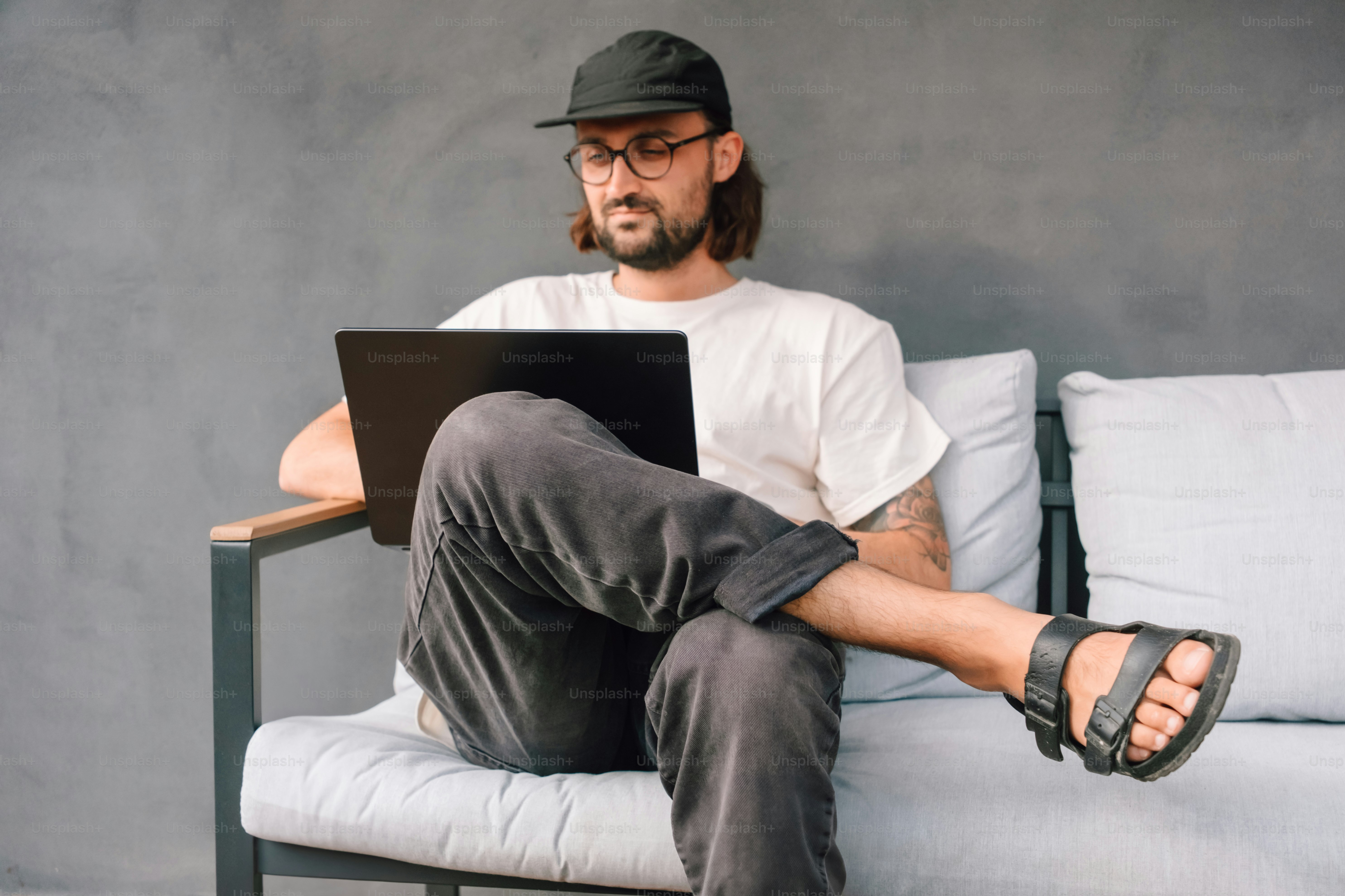Man wearing baseball cap and glasses using laptop on couch.